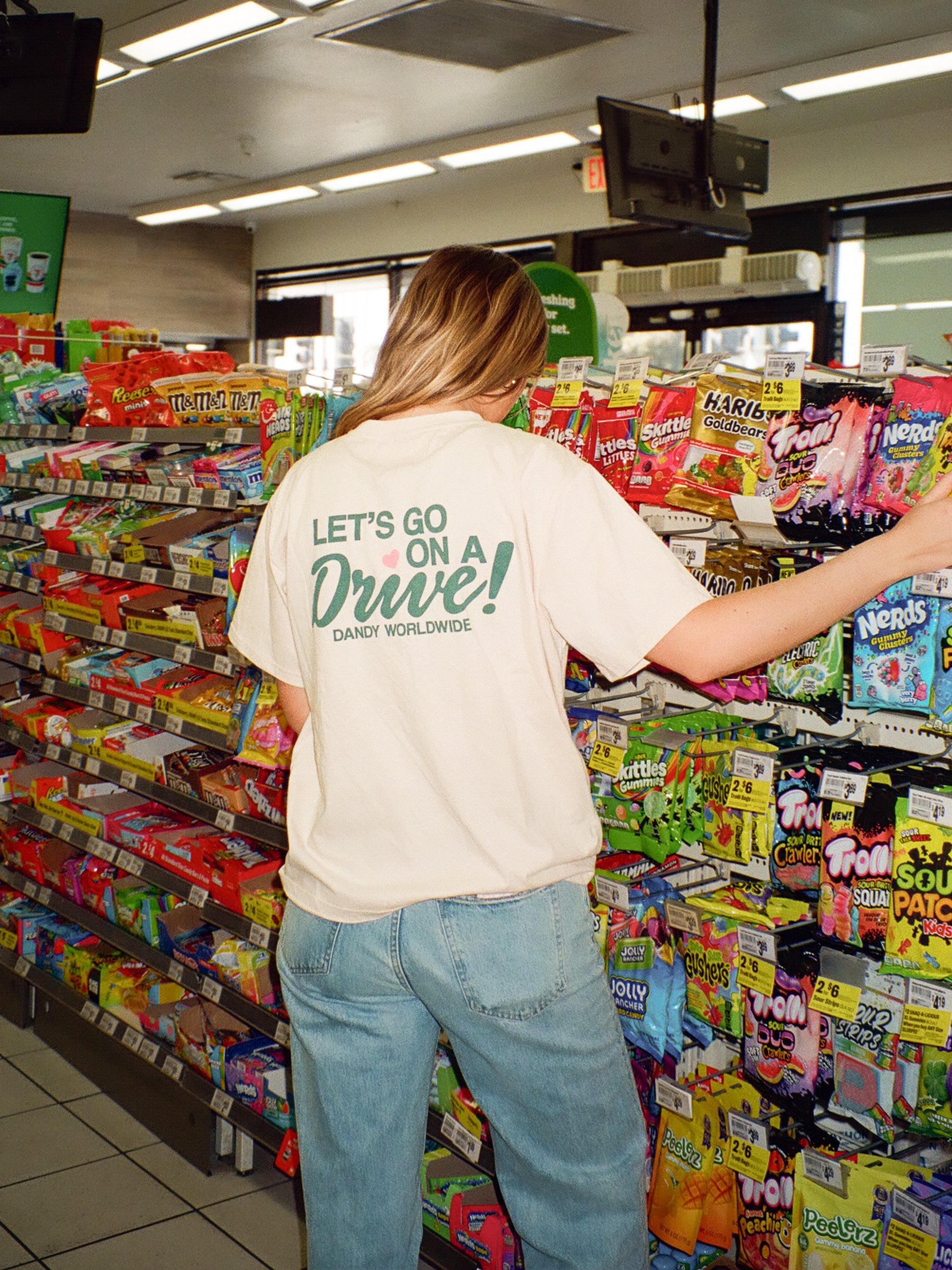 Person shopping in a candy aisle with colorful packaging. Shirt reads LETS GO ON A DRIVE
