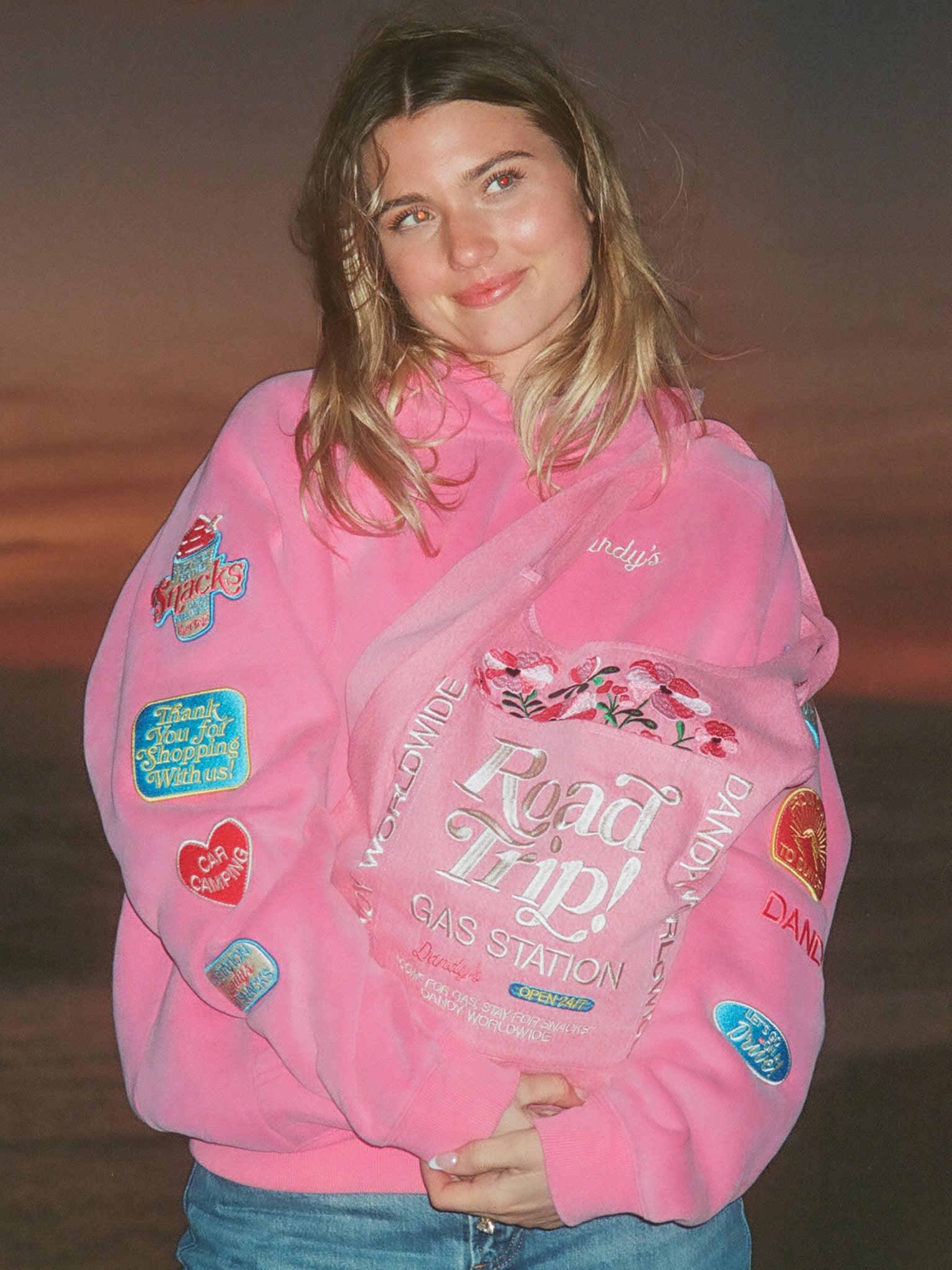 Person wearing a pink hoodie with various patches and text, standing against a sunset sky.