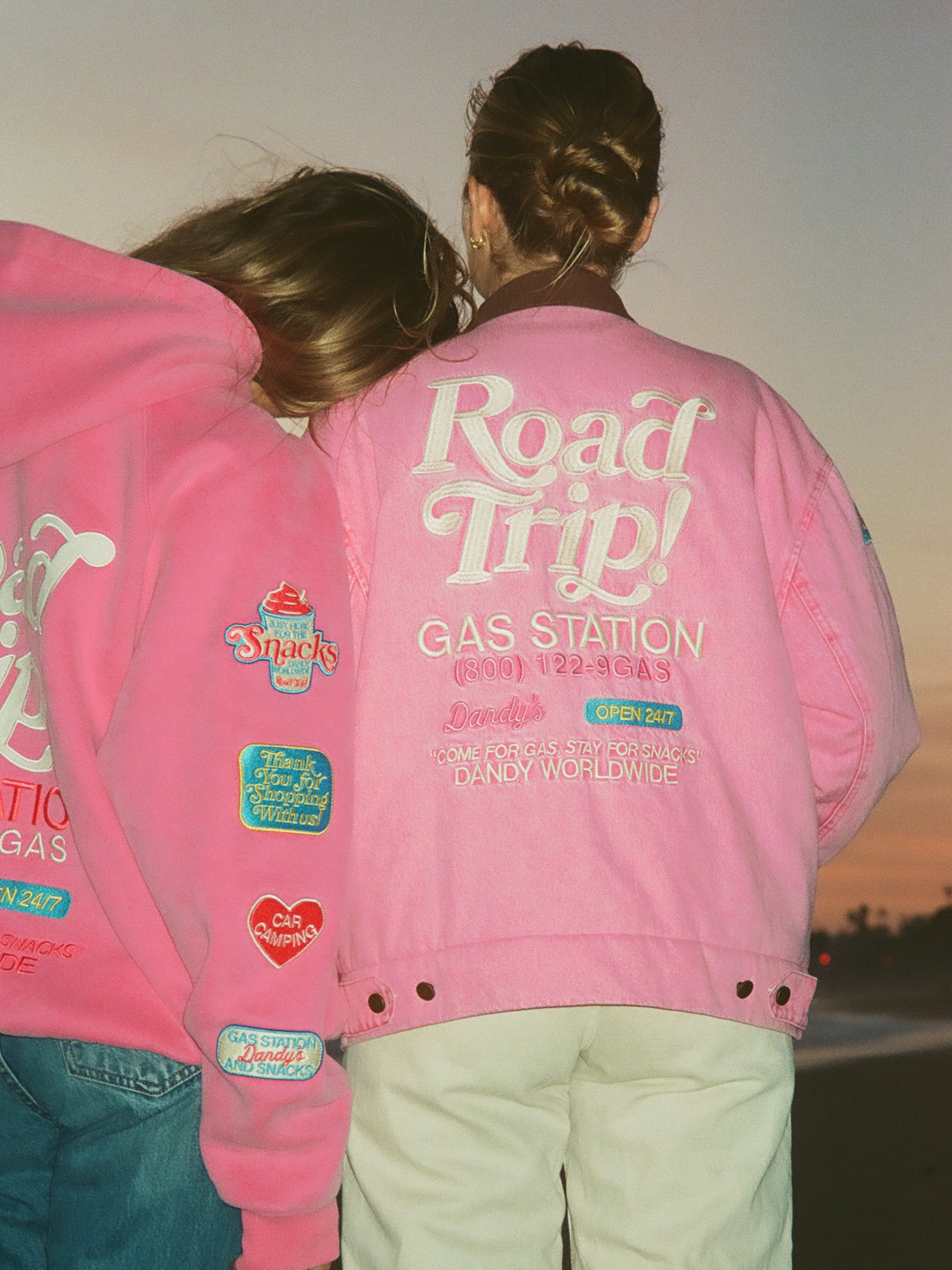 Two people wearing pink jackets with text and graphics that read "ROAD TRIP! GAS STATION", standing close together against a sunset background.