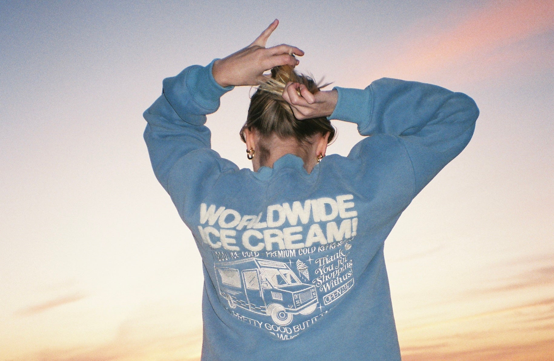 Person tying hair in blue hoodie with WORLDWIDE ICE CREAM on back, set against a pastel sunset sky.