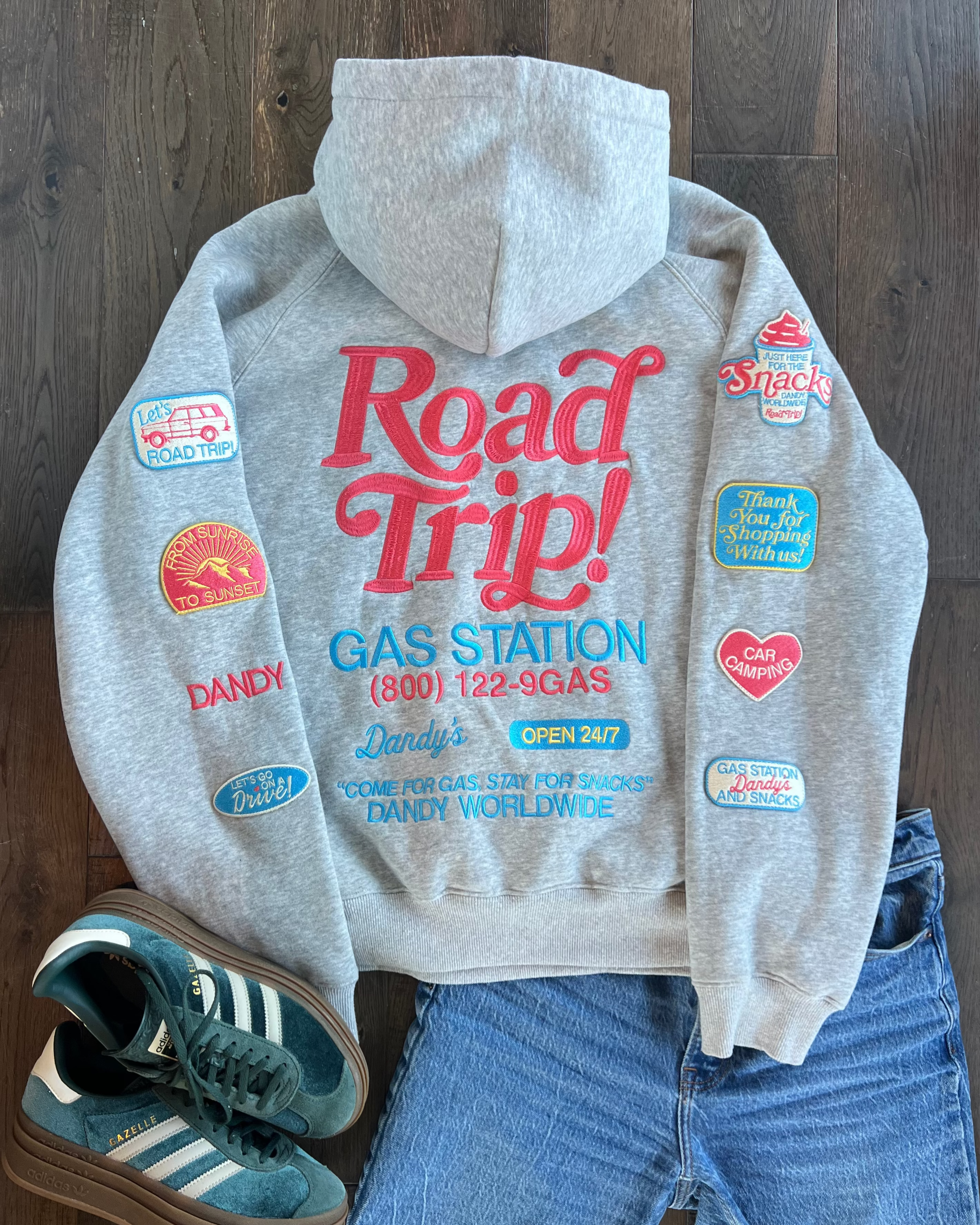 Gray hoodie with 'Road Trip!' text and patches, paired with blue jeans and sneakers on a wooden floor.