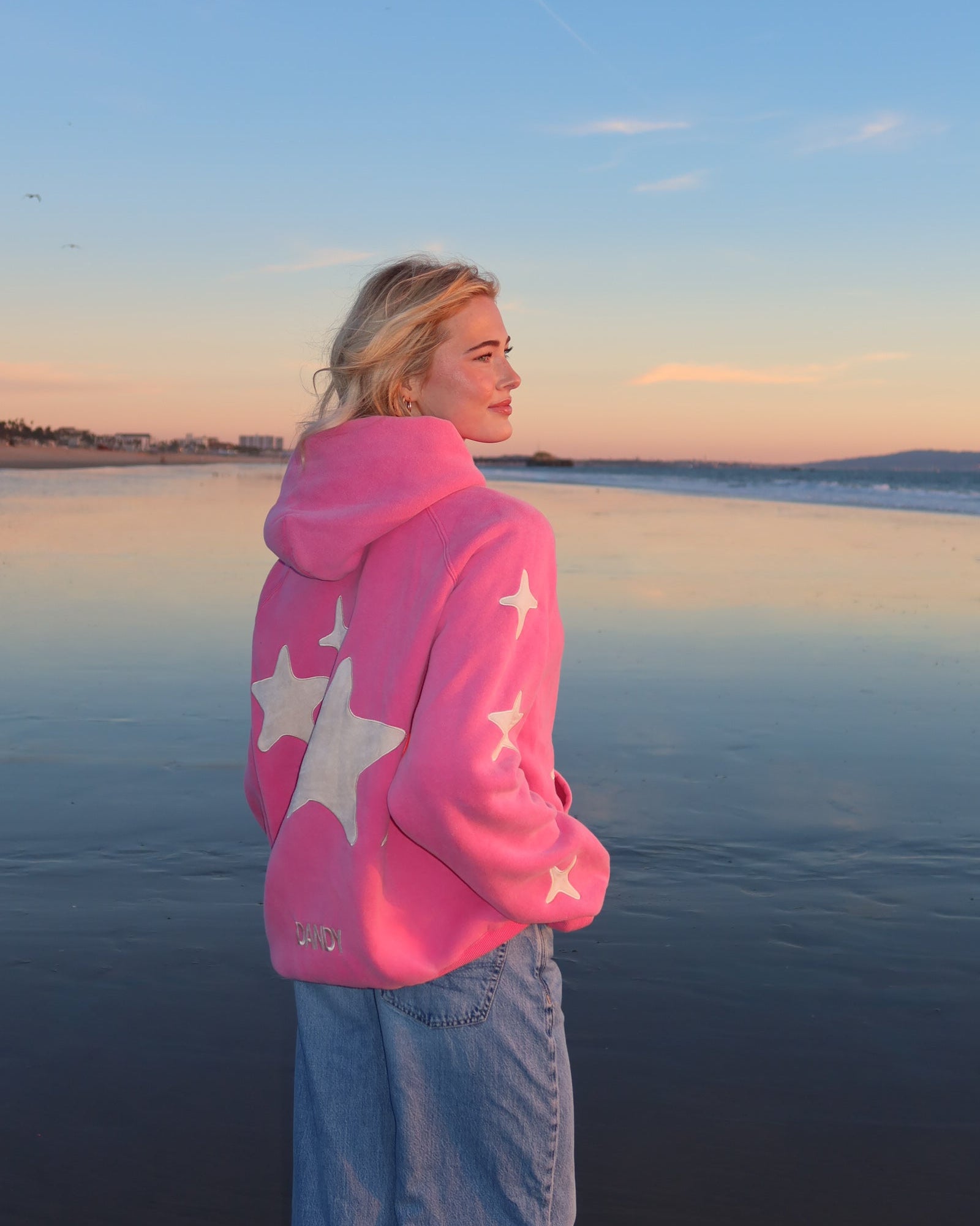 Person wearing a pink hoodie with star patterns by a body of water at sunset.