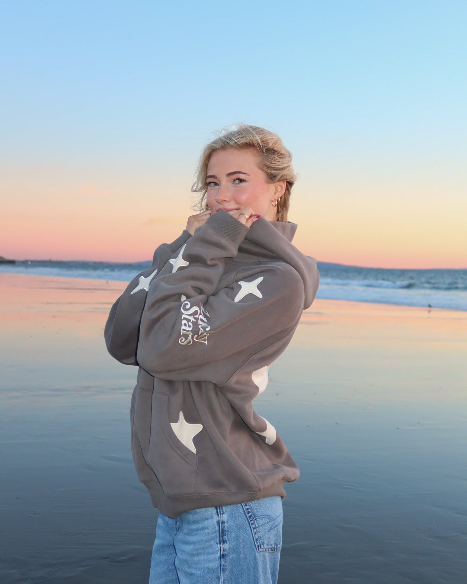 Blonde girl looking at the camera and standing on a beach at sunset while wearing a gray star hoodie