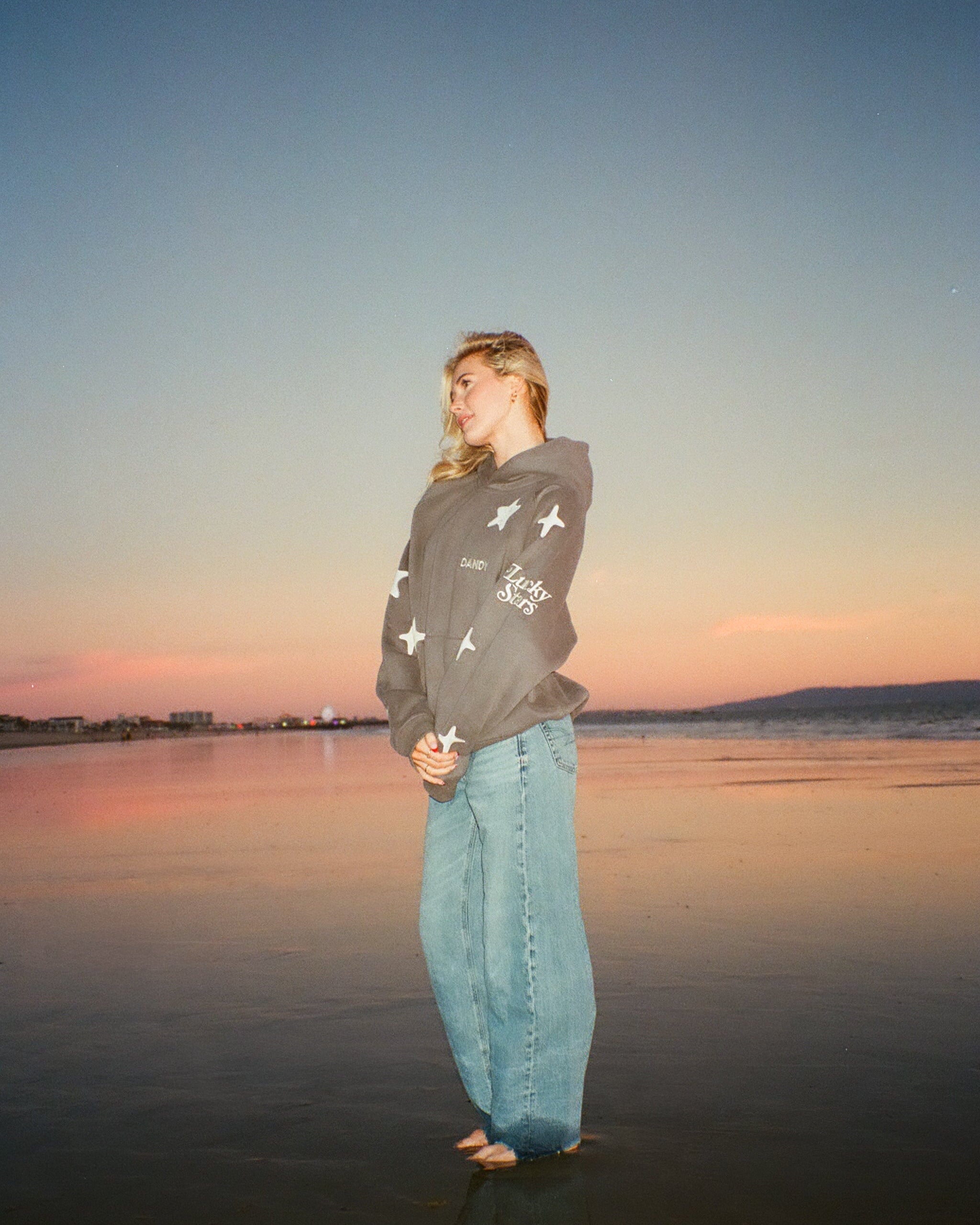 Blonde girl wearing a gray star hoodie on the beach at sunset