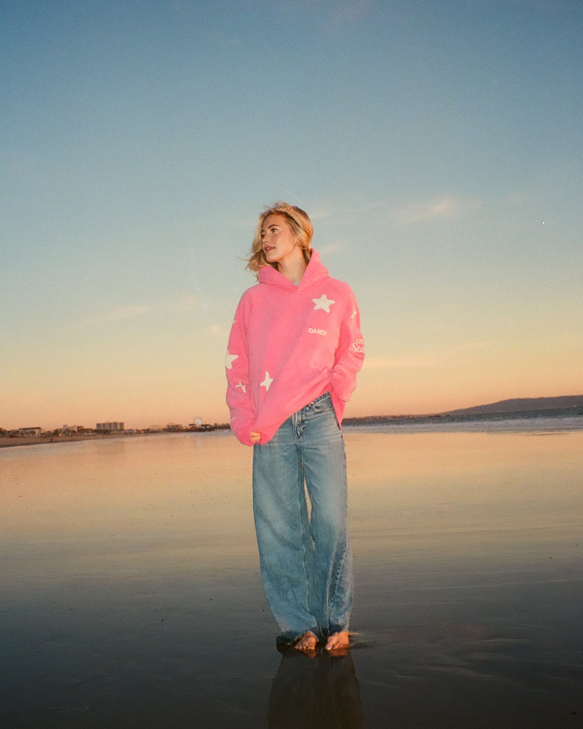 Person wearing a pink hoodie and jeans standing on a beach at sunset.