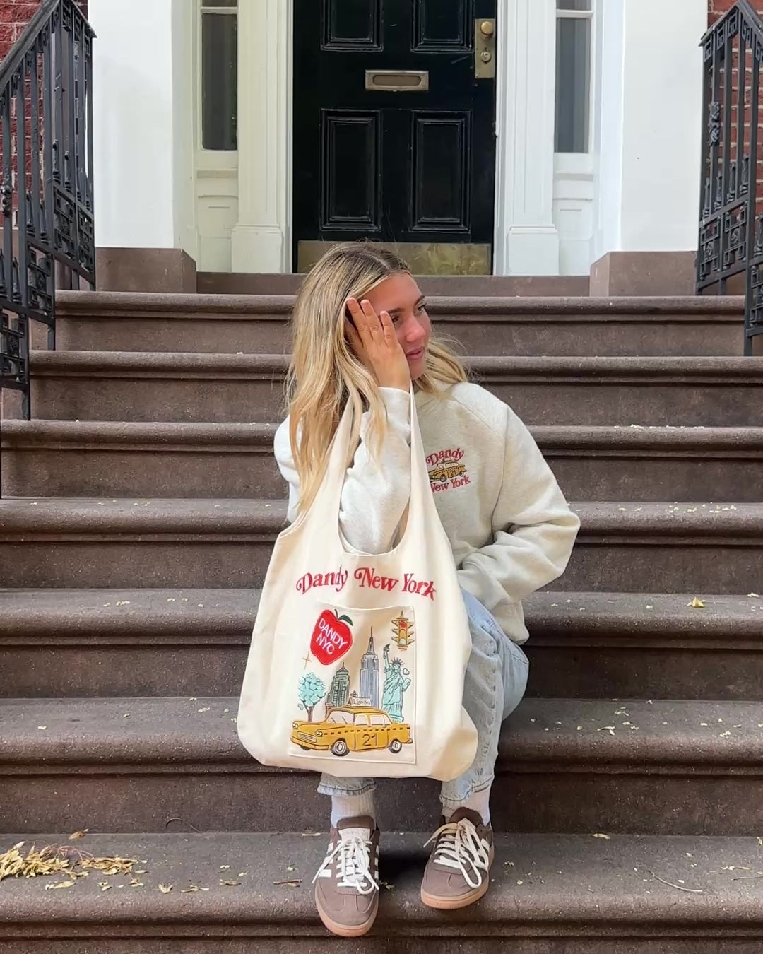 video of a girl sitting on stairs outside of an apartment in new york. she is holding a tote bag that says dandy new york, with a taxi, apple, buildings, and the statue of liberty, all on the pocket of the tote bag. tote bag is cream canvas.