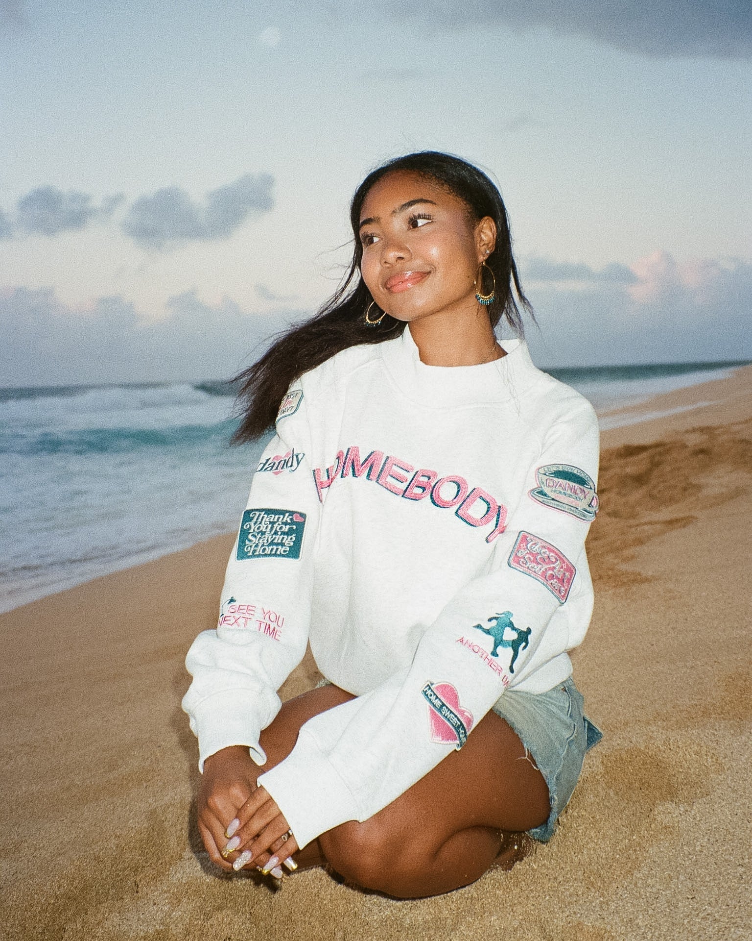 Woman sitting on a beach wearing a white sweatshirt with colorful graphics.