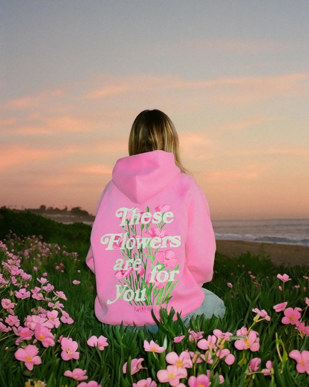 Person wearing a pink hoodie with text, standing in a field of flowers with a sunset in the background.
