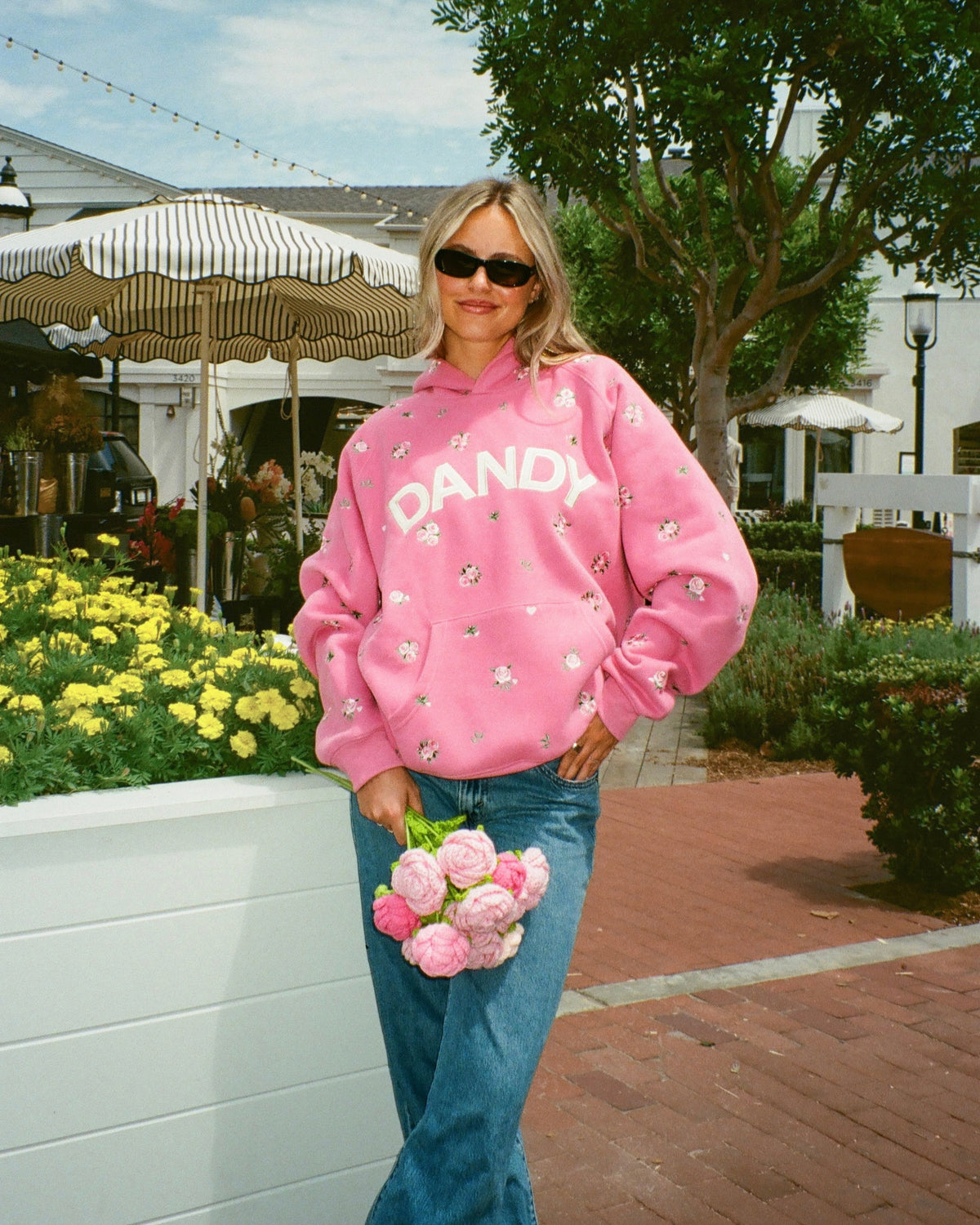 A woman wearing a pink pullover hoodie with 'DANDI' printed across the chest, styled with blue jeans and holding a bouquet of pink flowers in an outdoor patio setting.
