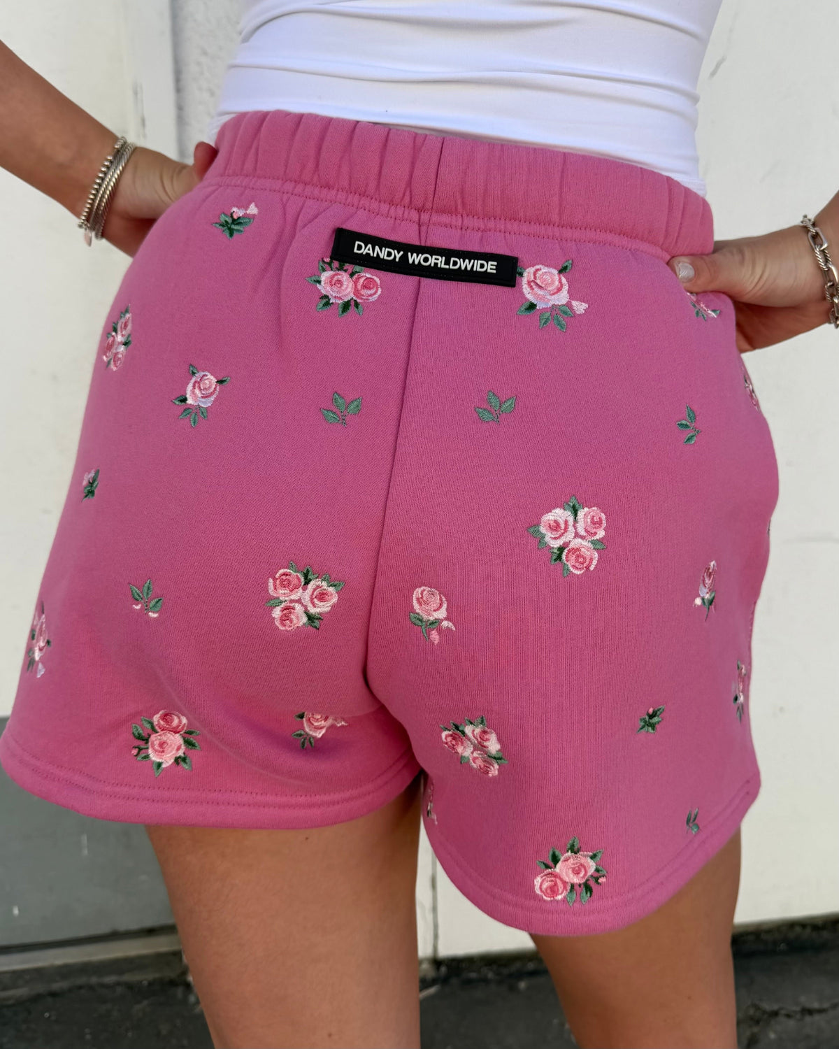 Pink sweat shorts with a floral rose pattern, featuring a black brand label that reads 'DANDY WORLDWIDE' on the back waistband, worn by a model.