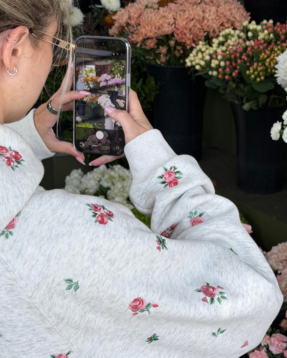 A person wearing a light gray heathered sweatshirt with small pink rose and green leaf embroidery, holding a smartphone to take a photo in a flower shop setting.