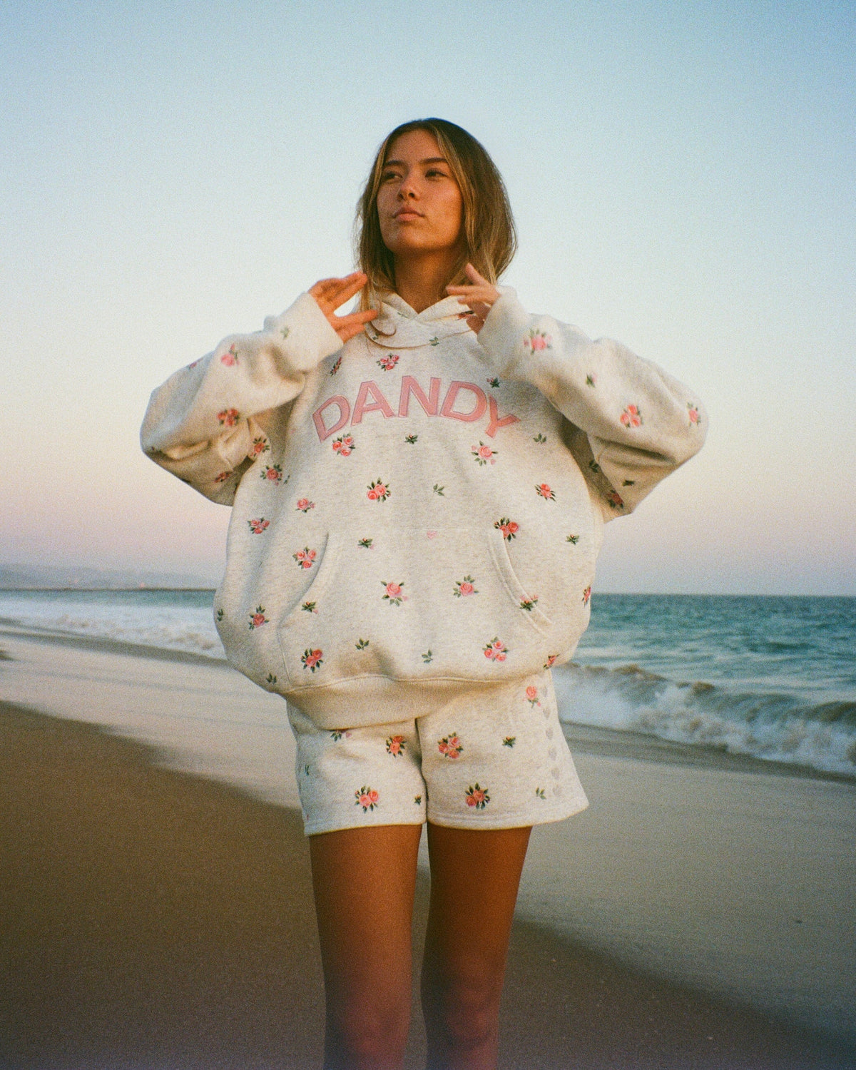 A woman stands on a beach wearing a cream-colored matching hoodie and shorts set with a small pink and green floral pattern and pink 'DANDY' text on the front.