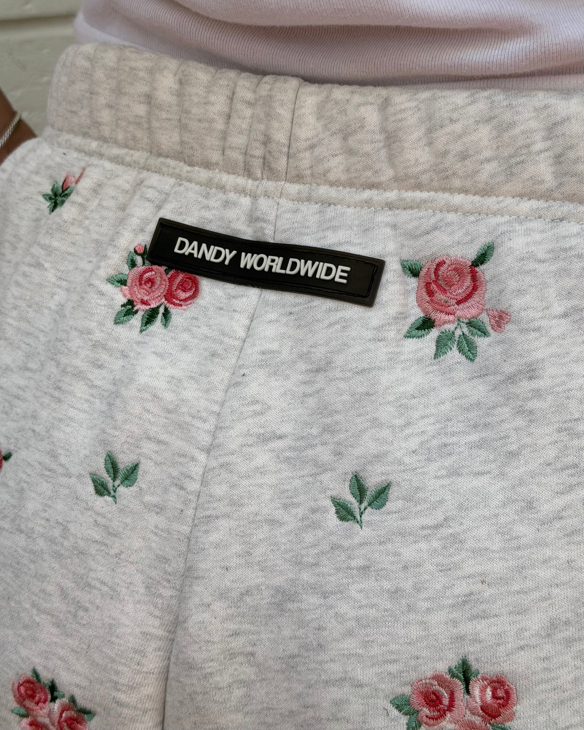 Close-up of a pair of light heather gray sweatpants with embroidered pink roses and green leaves, featuring a black brand patch that reads 'DANDY WORLDWIDE' at the waistband.