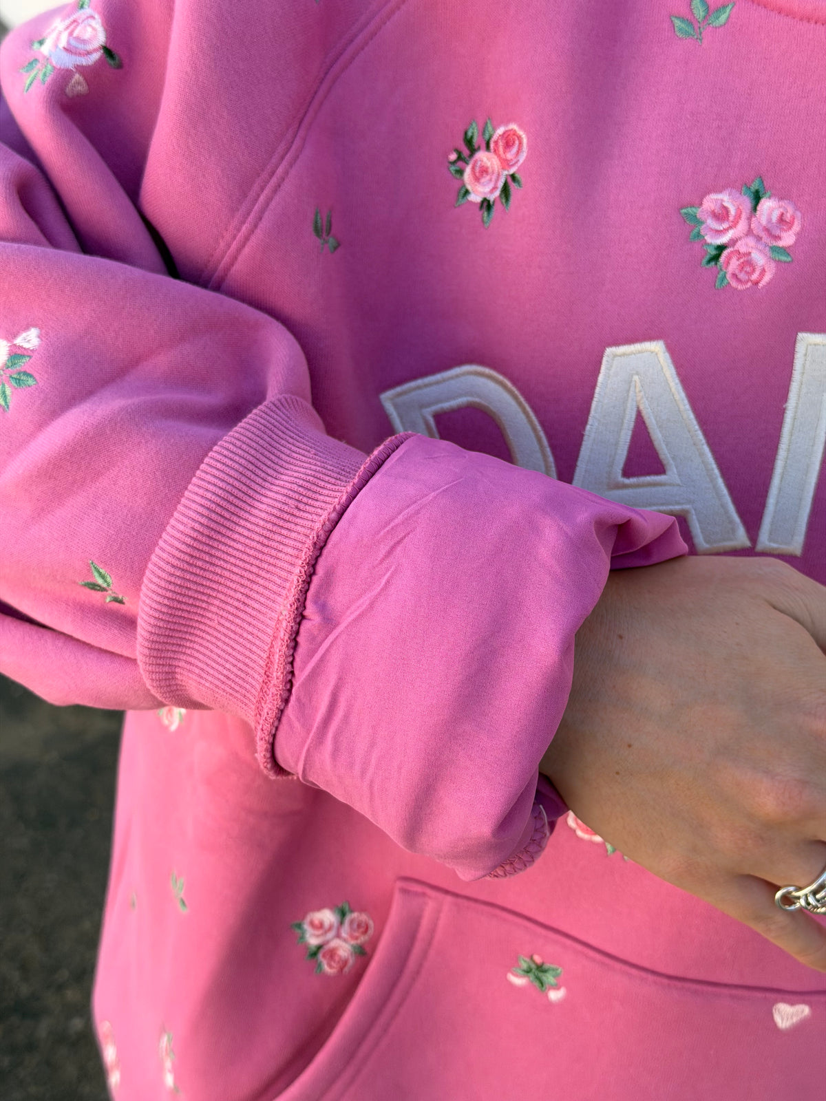 A close-up of a person wearing a pink hoodie with embroidered pink roses and white lettering on the chest.
