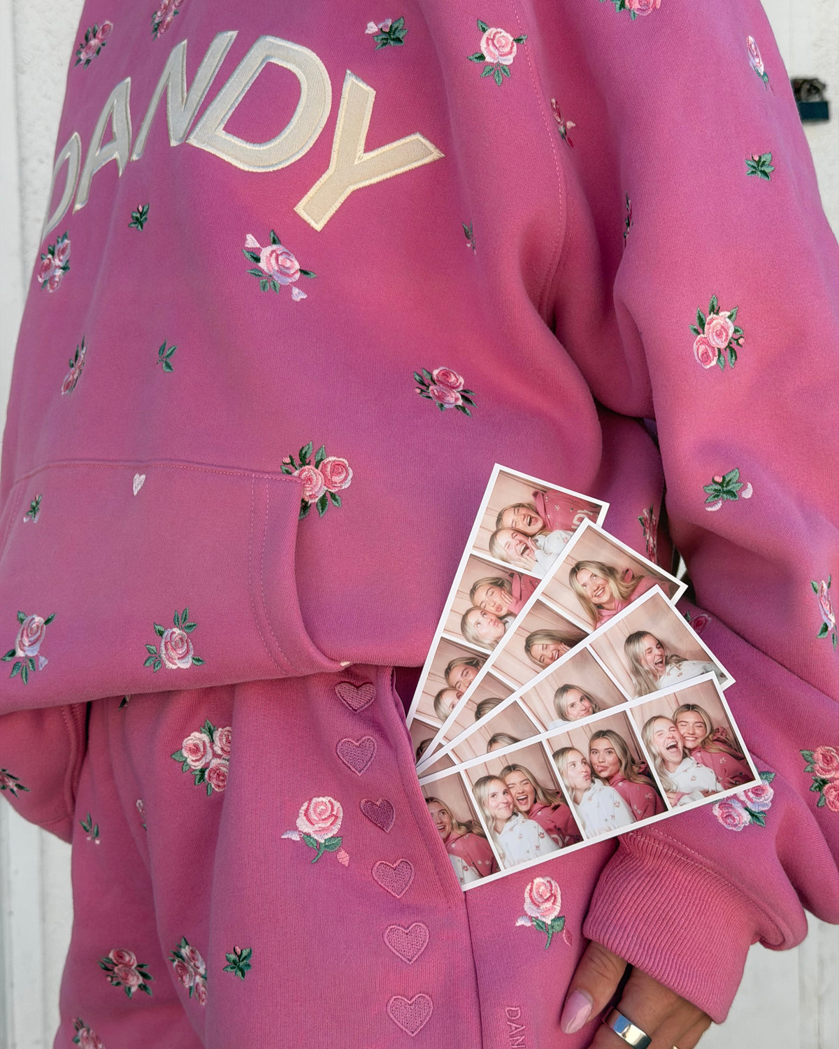 A close-up of a person wearing a pink sweatshirt and matching sweatpants. The sweatshirt has a large gold 'DANDY' text print on the chest and is decorated with small pink rose and heart motifs.