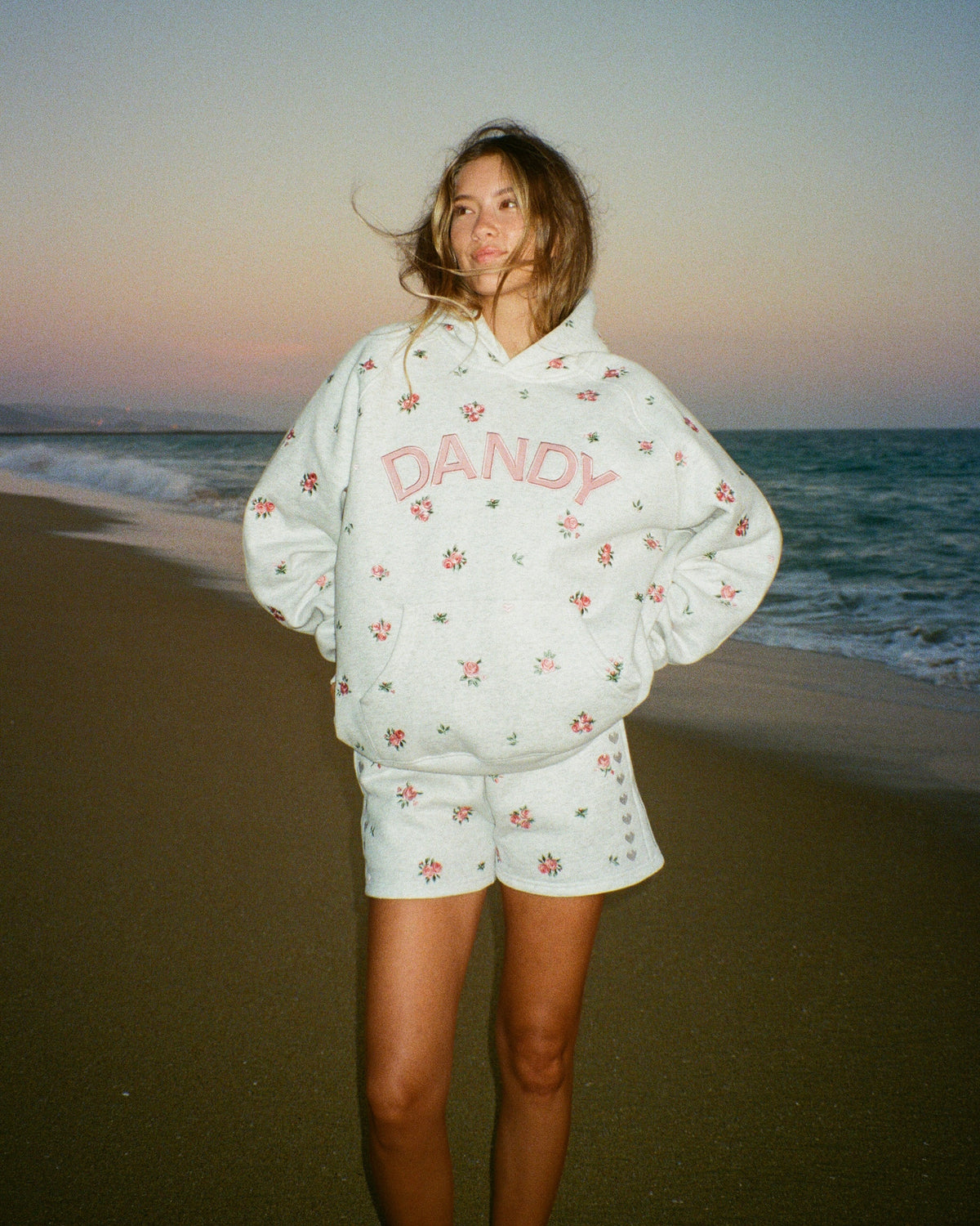 A woman stands on a beach wearing a white oversized hoodie and matching shorts set, both covered in a small pink floral print, with the word 'DANDY' printed in pink across the chest. The background shows the ocean and a sunset sky.