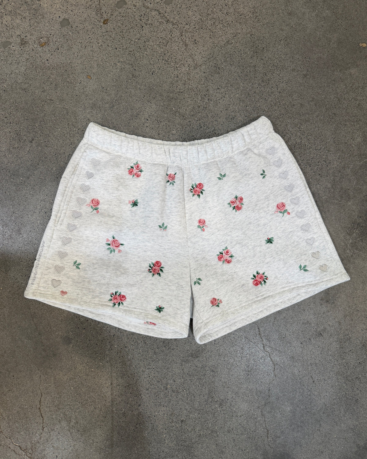 White textured gray shorts with small pink and green floral embroidery, laid flat on a gray concrete floor.