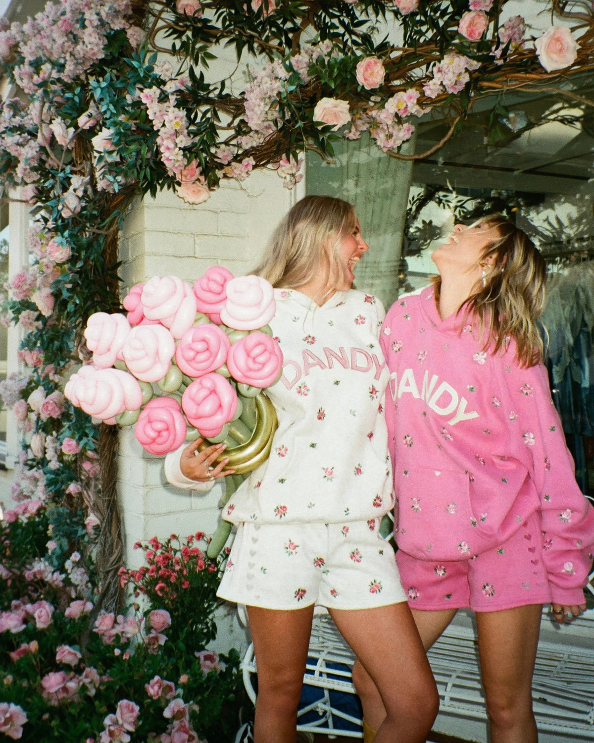 Two women posing in matching floral-print lounge sets, one in white and one in pink, holding oversized pink rose props in front of a white brick wall decorated with pink flowers.