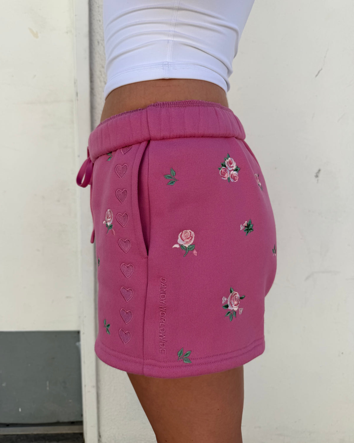A person wearing pink drawstring shorts with embroidered pink roses and green leaves, paired with a white ribbed top, standing against a light off-white wall.