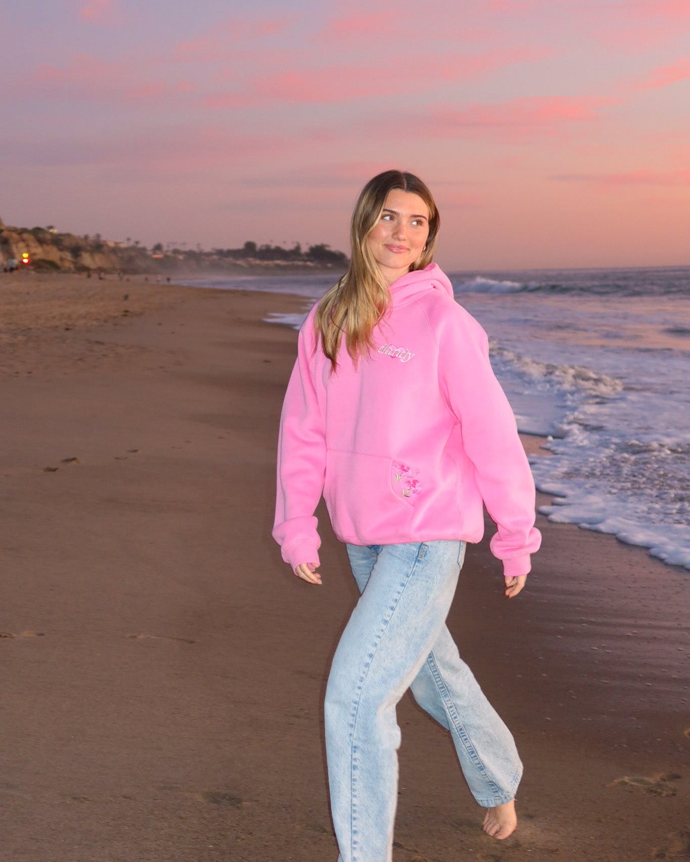Person wearing a pink hoodie and light blue jeans standing on a beach at sunset.