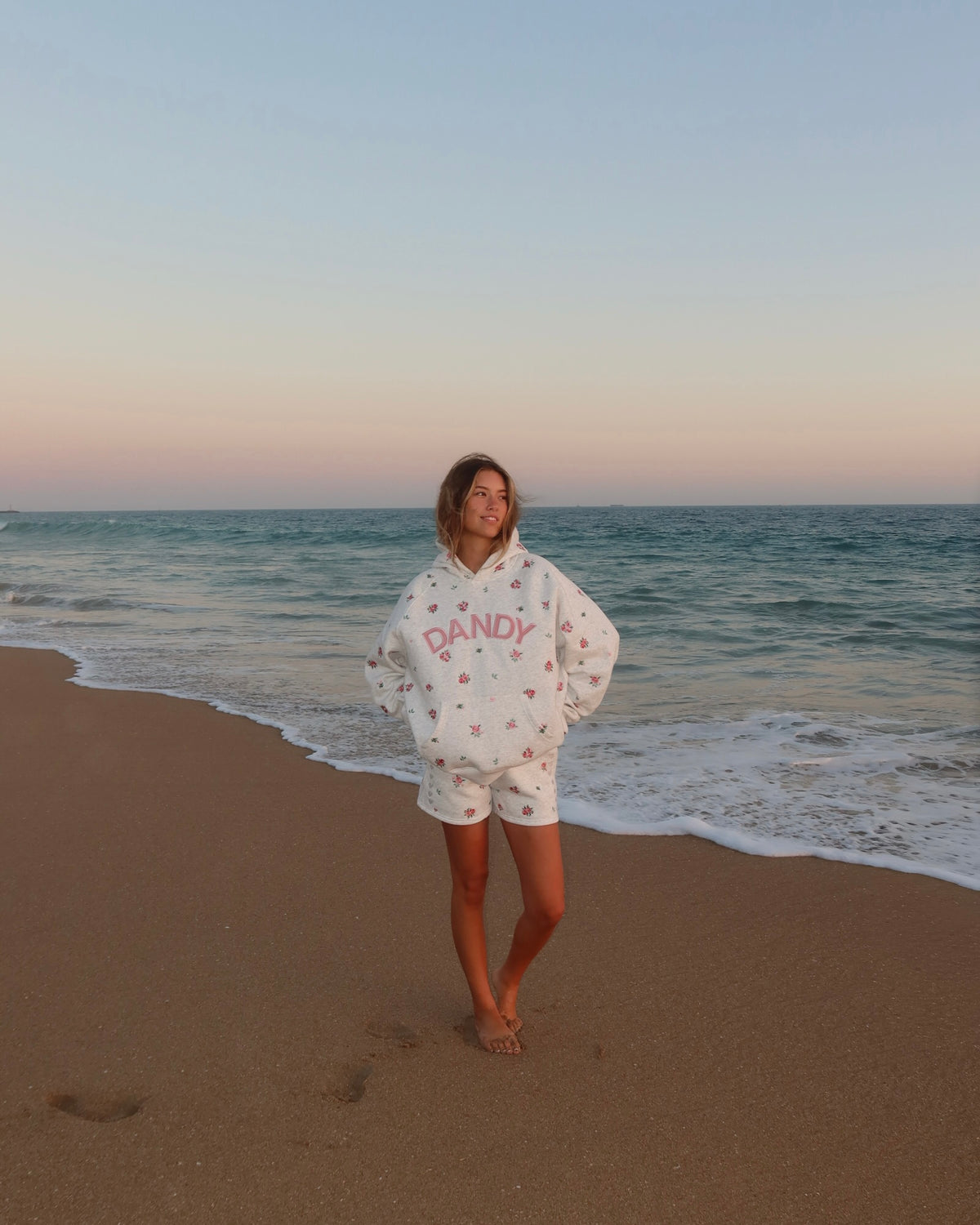 A woman stands on a sandy beach at sunset wearing a white oversized hoodie and matching shorts with a small pink floral pattern and the word 'DANDY' printed in pink across the front.
