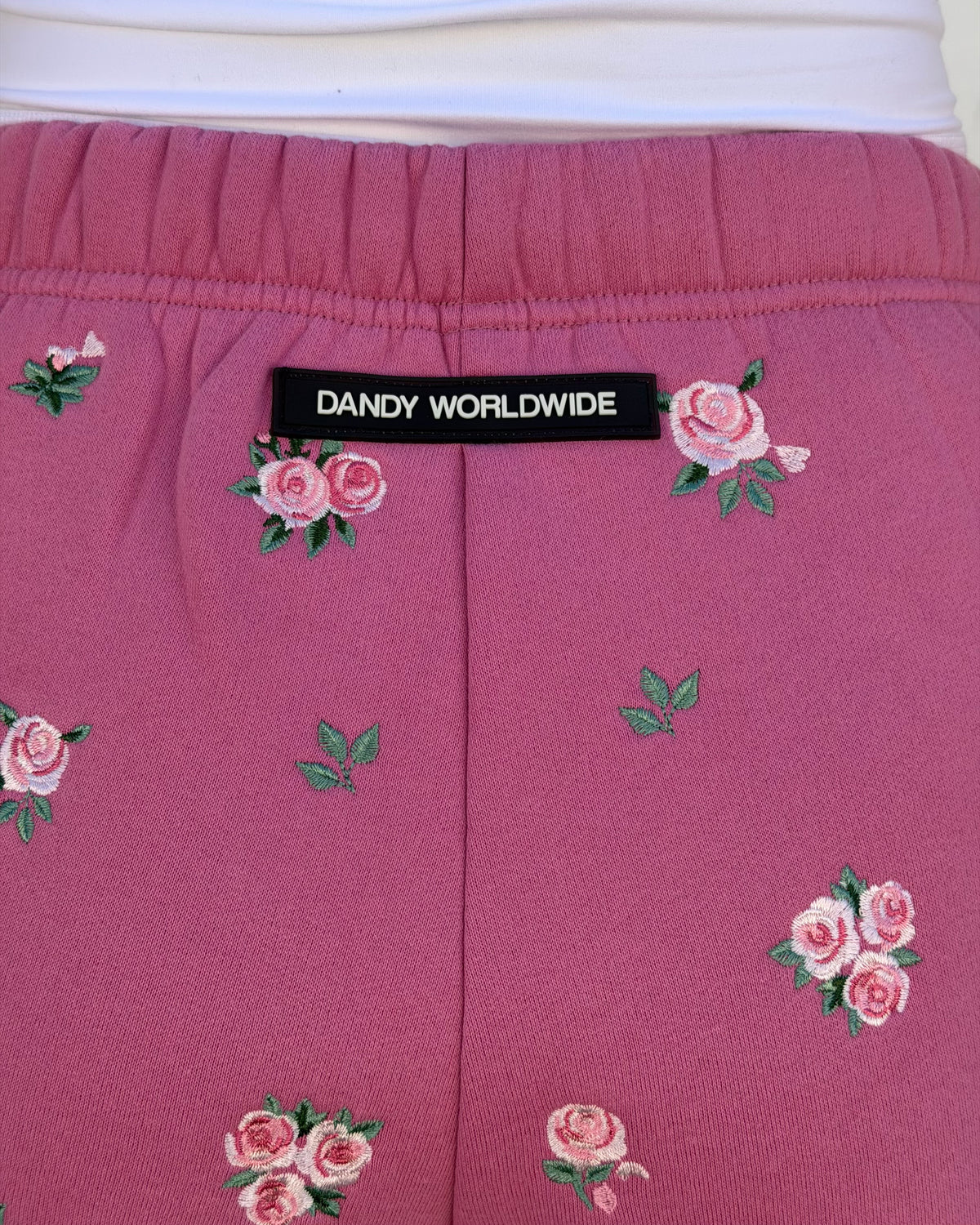 Close-up of pink embroidered pants with a black Dandy Worldwide label at the waistband, featuring pink rose and green leaf motifs.