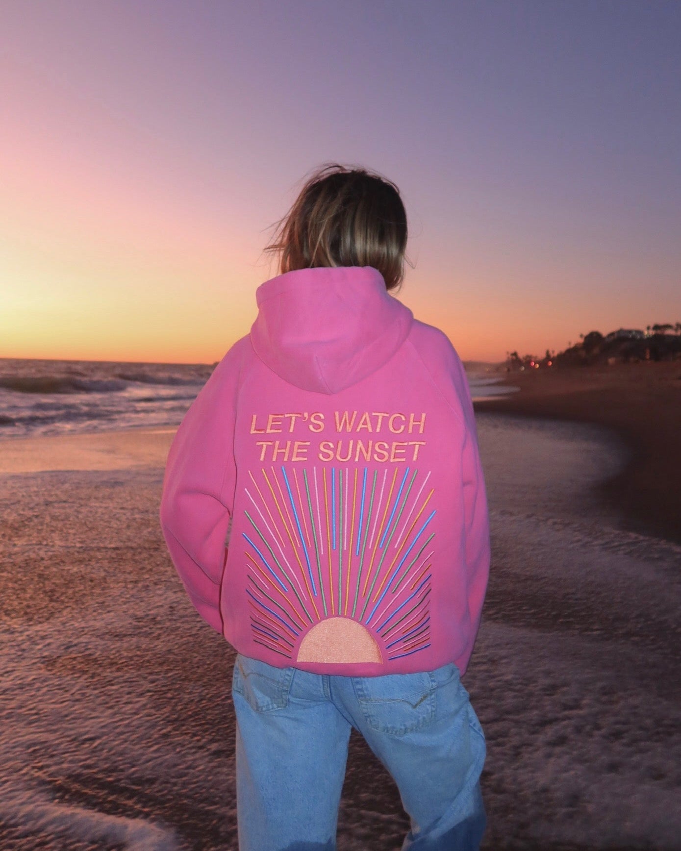 Person wearing a pink hoodie with text on the beach at sunset