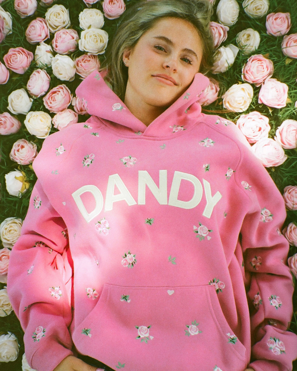 A woman lying down surrounded by pink and white roses, wearing a bright pink hoodie with 'DANDY' printed in white across the chest and small pink floral details.