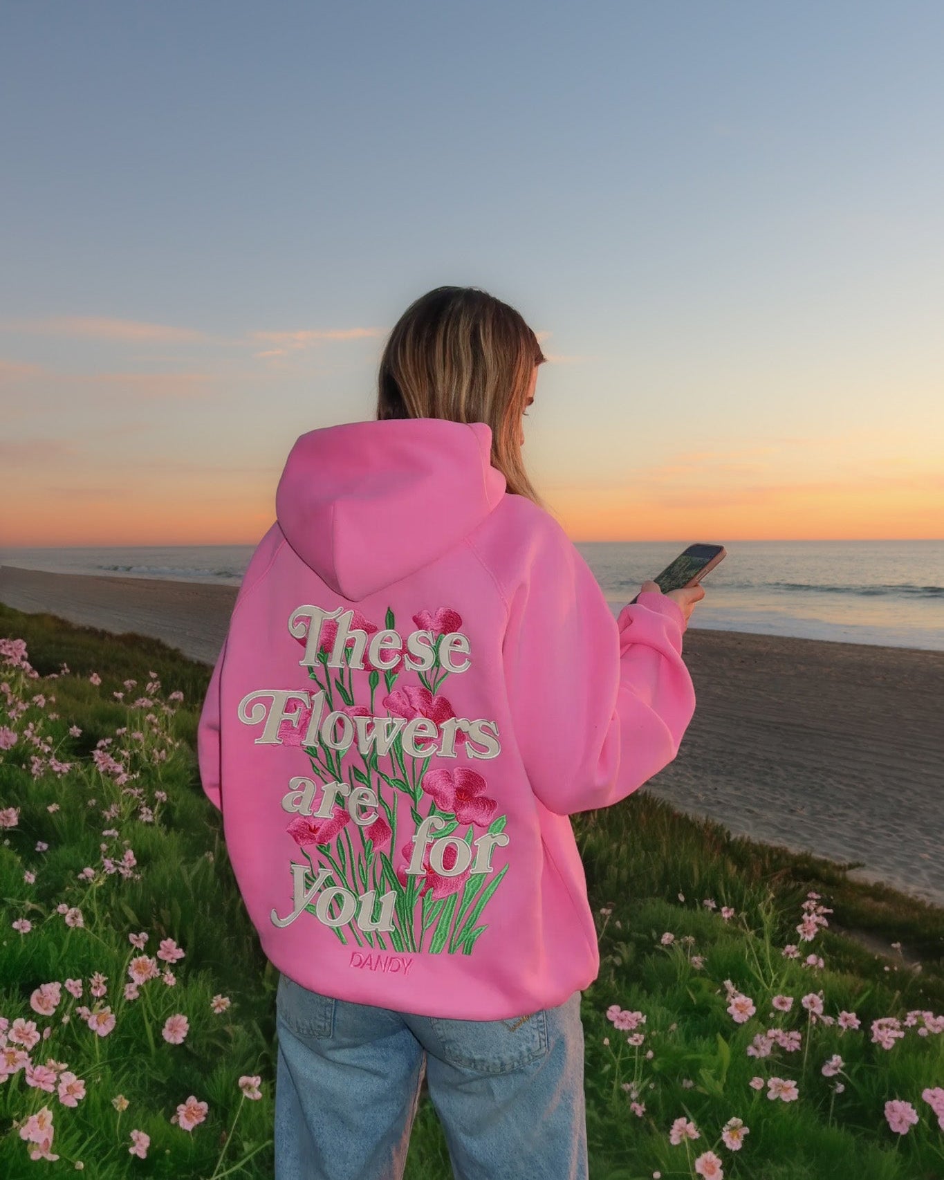 Person wearing a pink hoodie with a floral design and text, standing in a field of flowers with a sunset in the background.
