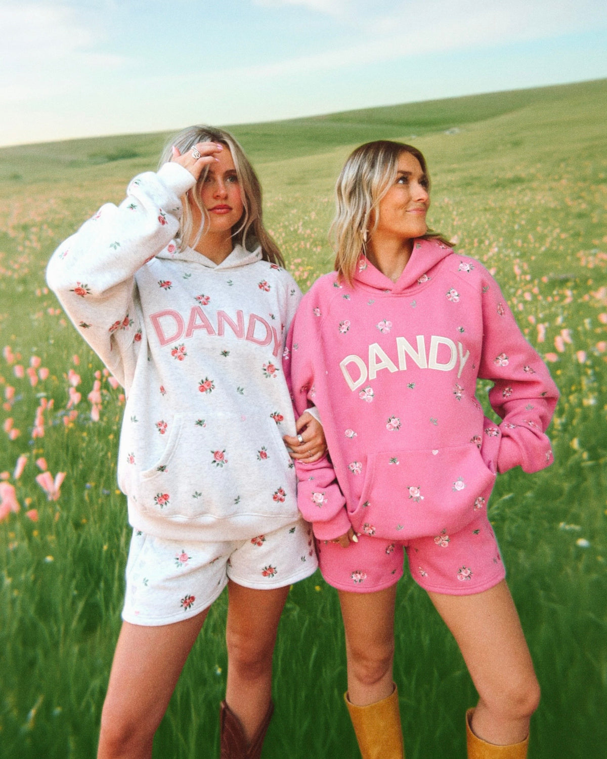 Two women standing in a grassy field wearing matching oversized hoodies and shorts sets, one in white and one in pink, both with floral prints and the word 'DANDY' across the front.