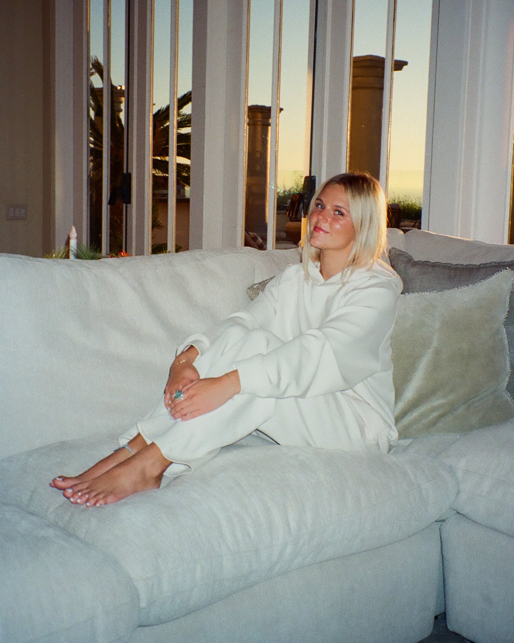 Woman in a white matching set sitting on a couch with a scenic view outside.