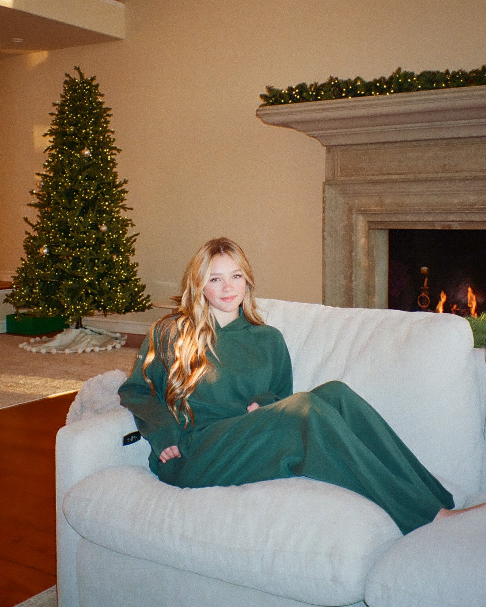 Woman in a green matching set sitting on a white couch in a living room with a Christmas tree and fireplace.