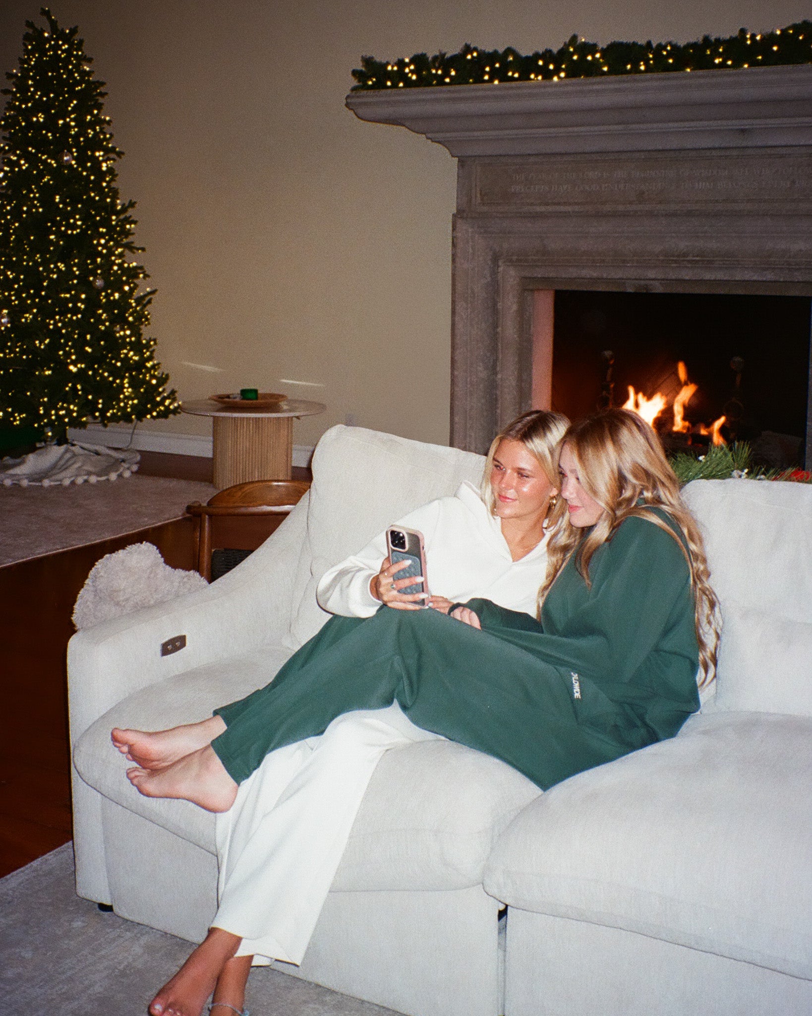 Two women sitting on a couch in a cozy living room with a Christmas tree and fireplace.