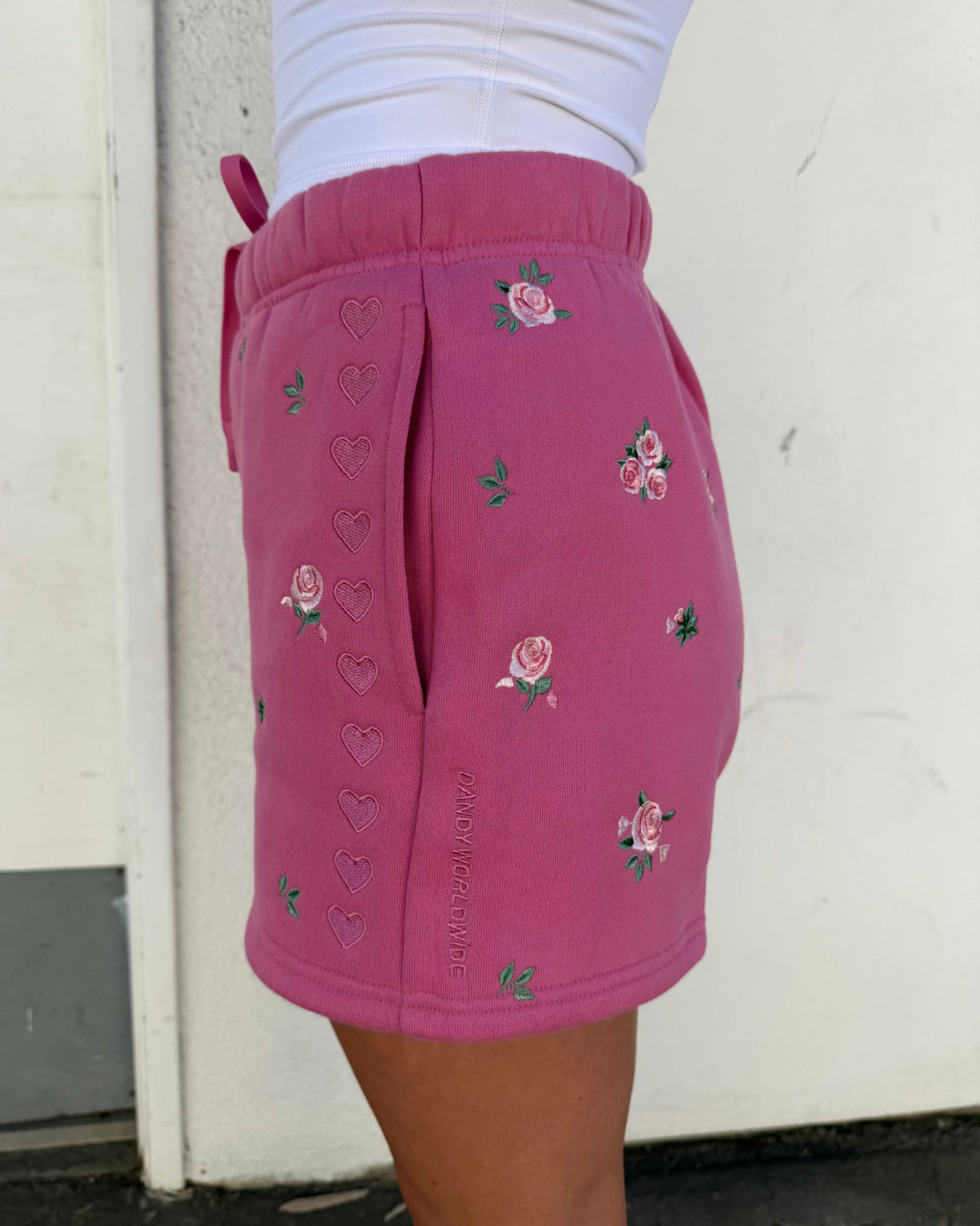 A person wearing bright pink shorts with embroidered pink roses and green leaves, plus tonal heart motifs, photographed outdoors against a light wall.