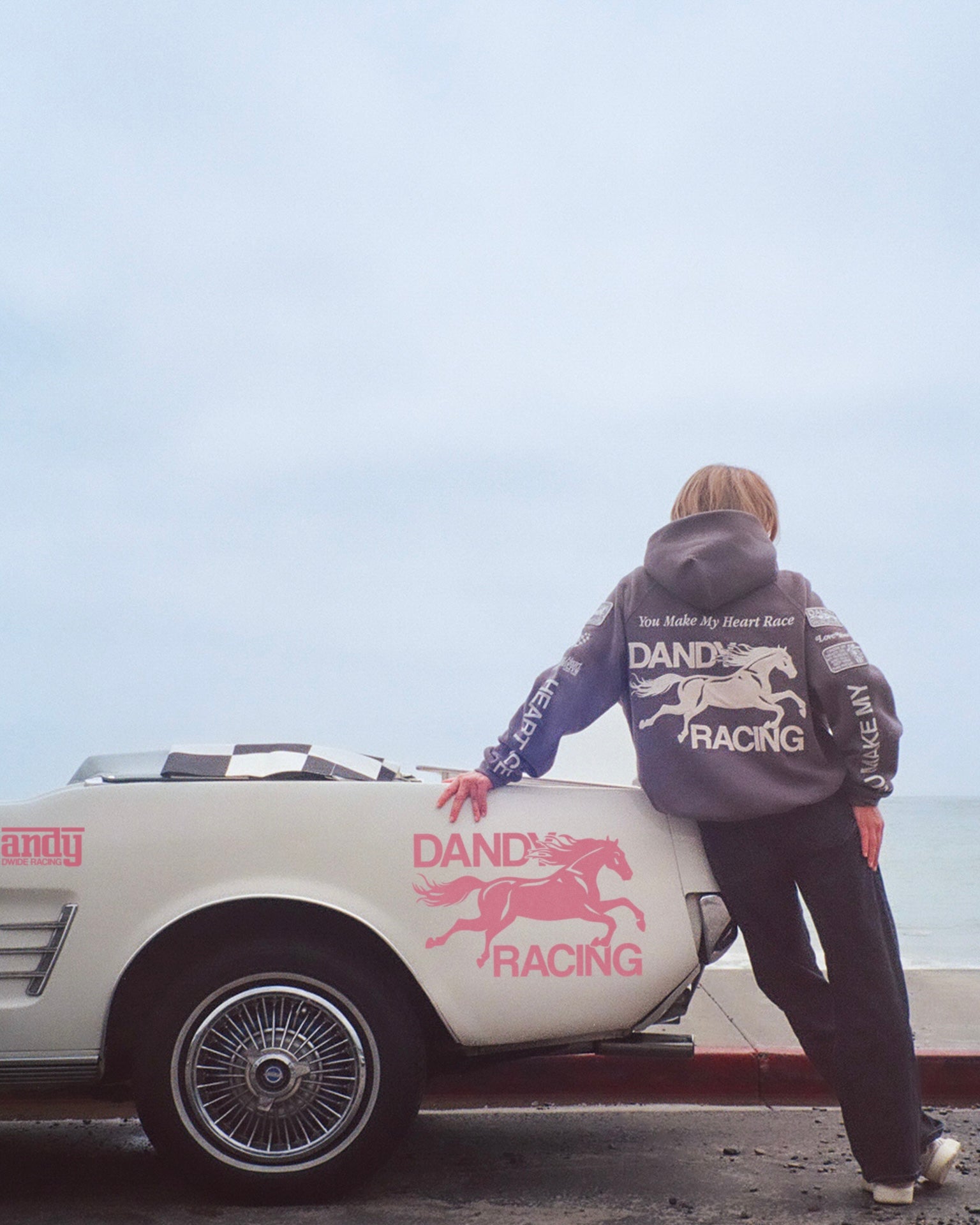 "Racing Heart" Oversized Lux Hoodie in Dark Gray leaning on a vintage car at the beach