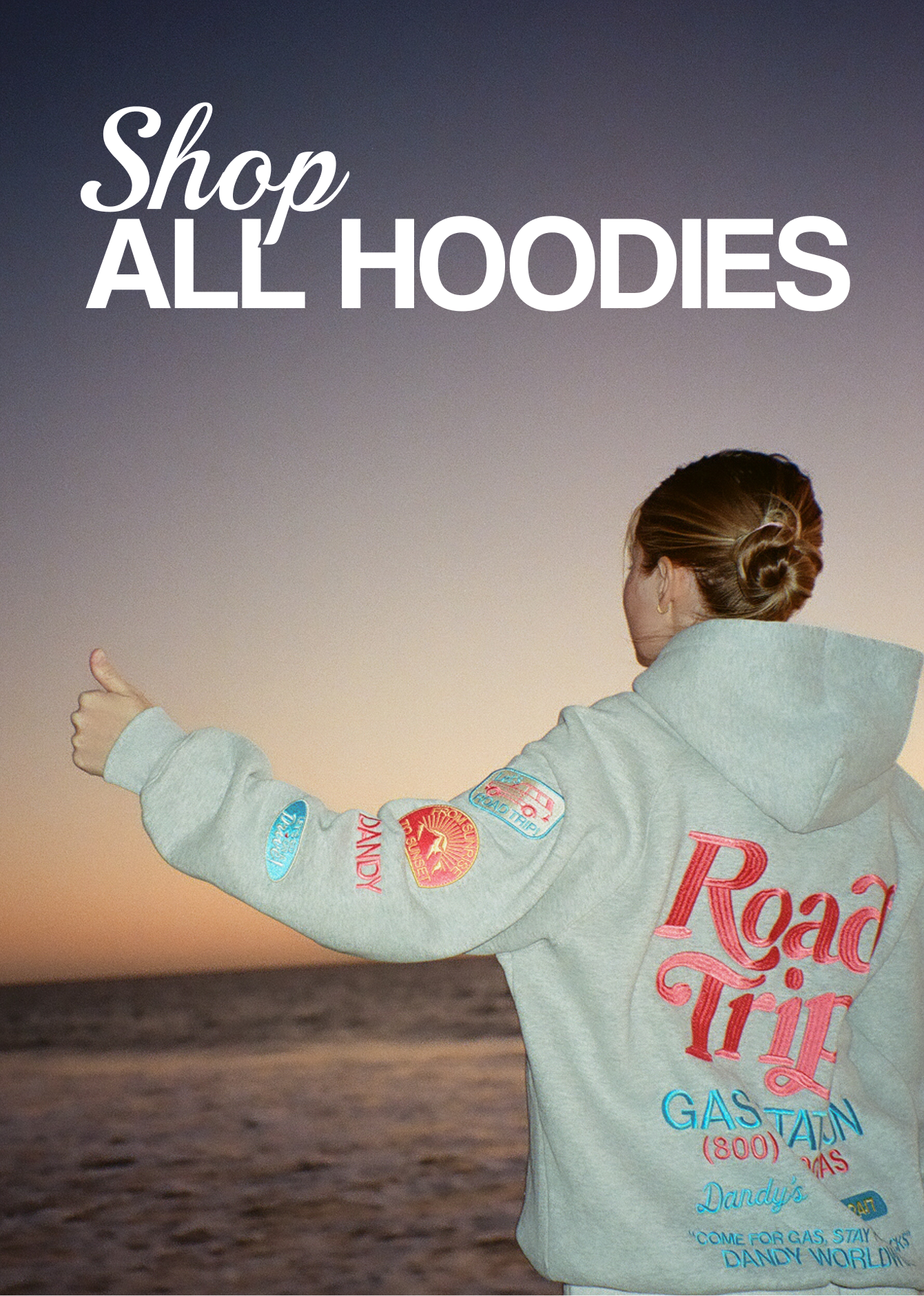 A young adult girl wearing a hoodie with 'Road Trip' text and patches, standing on a beach at sunset.