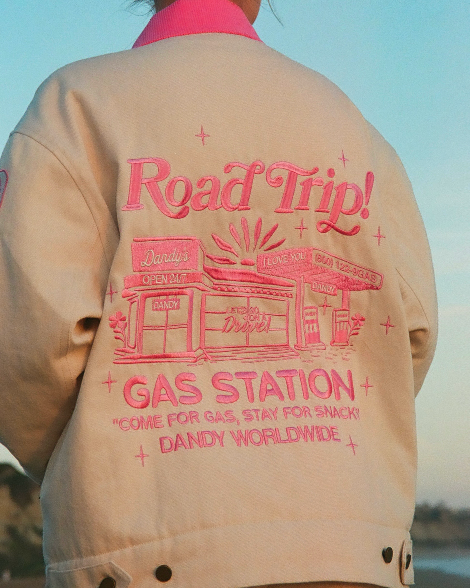 Beige jacket with pink 'Road Trip! Gas Station' text and graphics on a blurred natural background