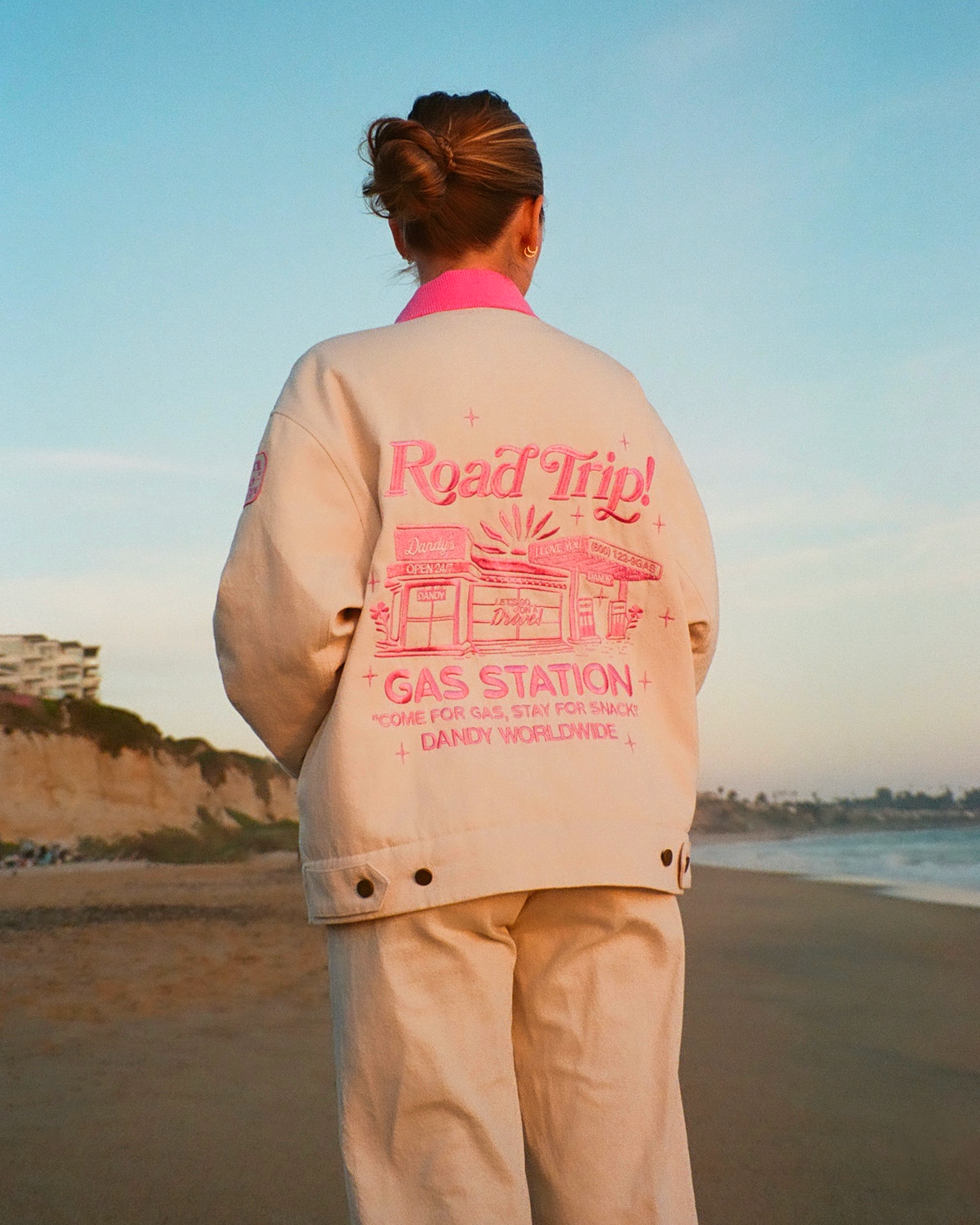 Person wearing a beige jacket with pink text on a beach that reads ROAD TRIP!