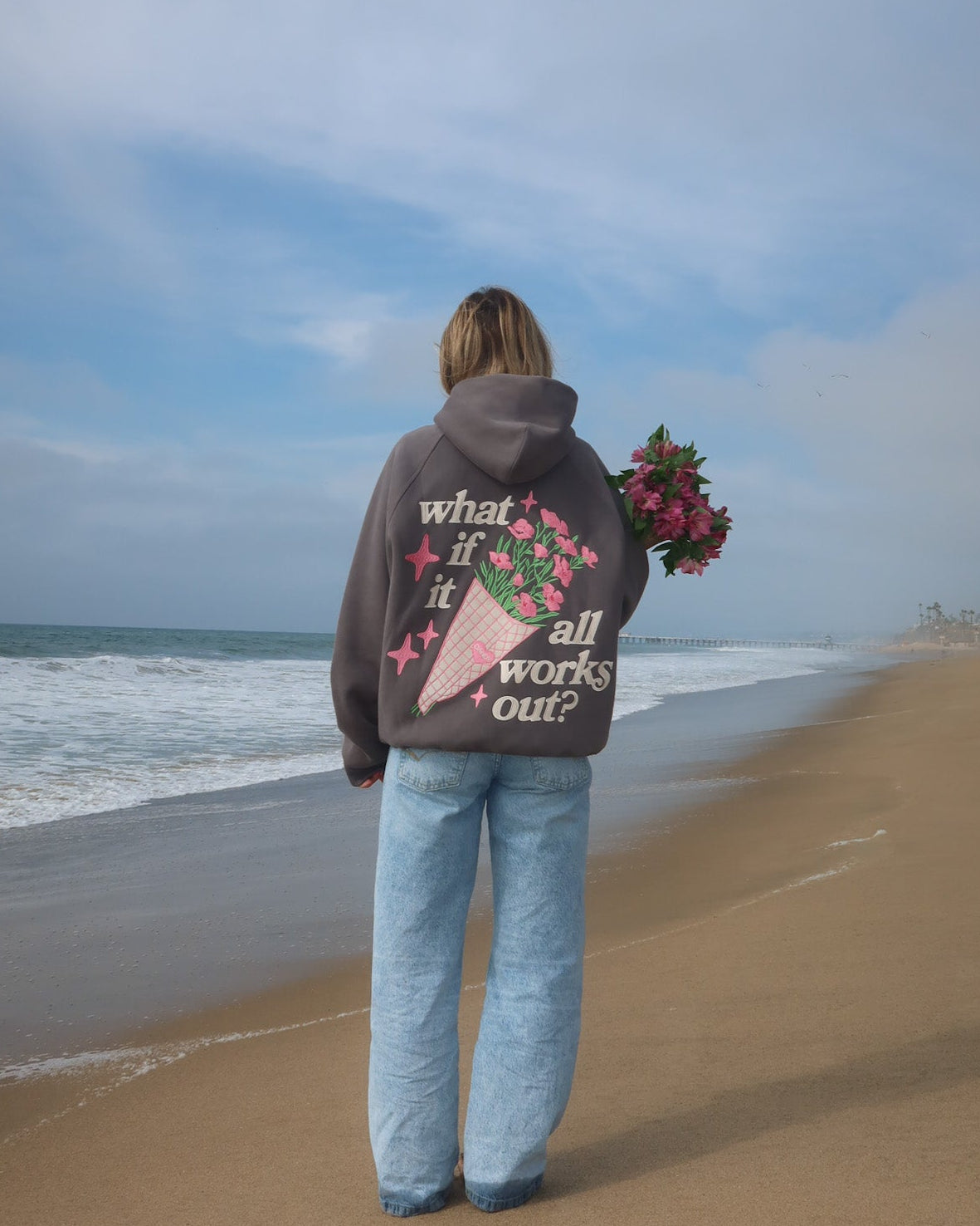 Person standing on a beach holding flowers, wearing a hoodie with text and graphics.