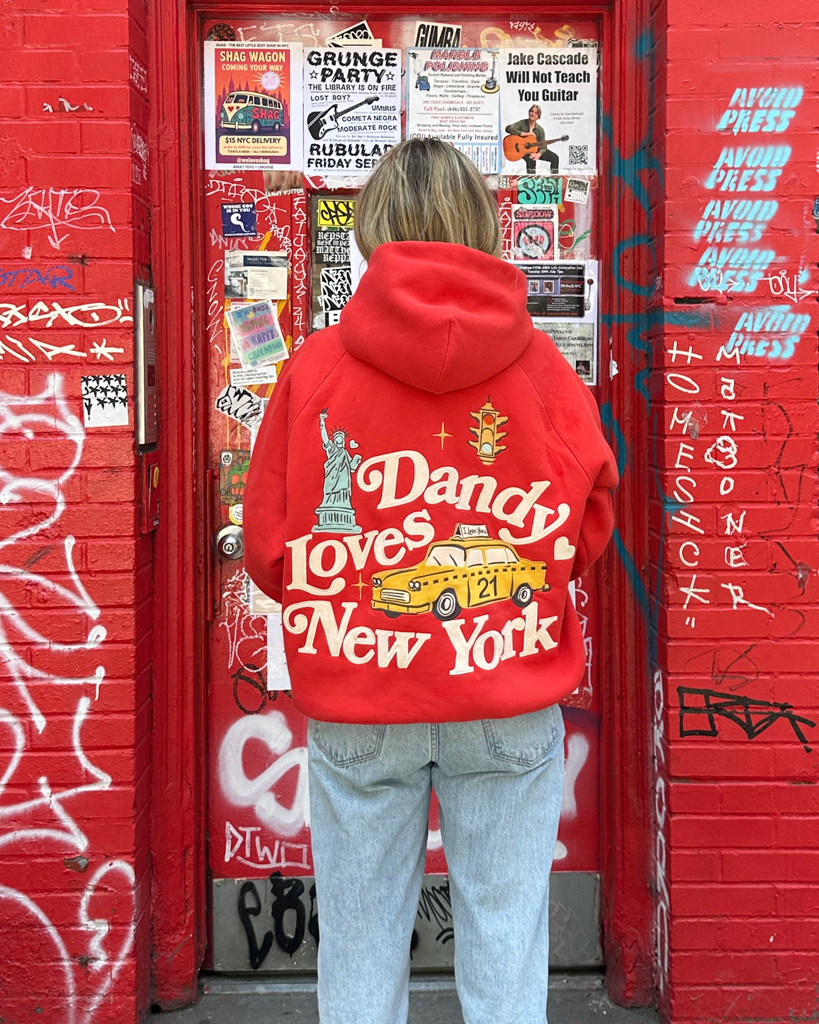 Person wearing a red hoodie with 'Dandy Loves New York' text in front of a graffiti-covered red door.