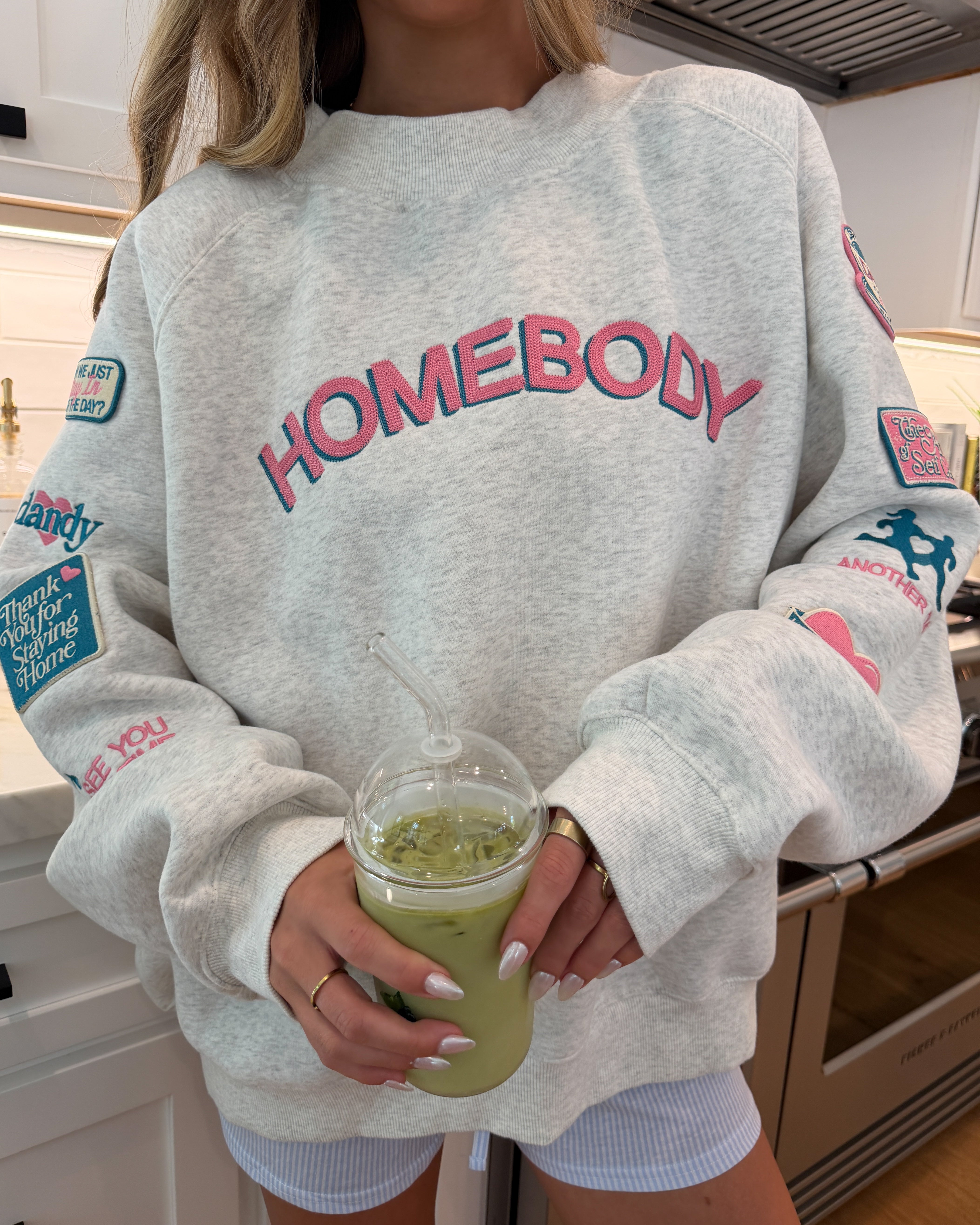 Person wearing a gray sweatshirt with 'HOMEBODY' text, holding a green drink.