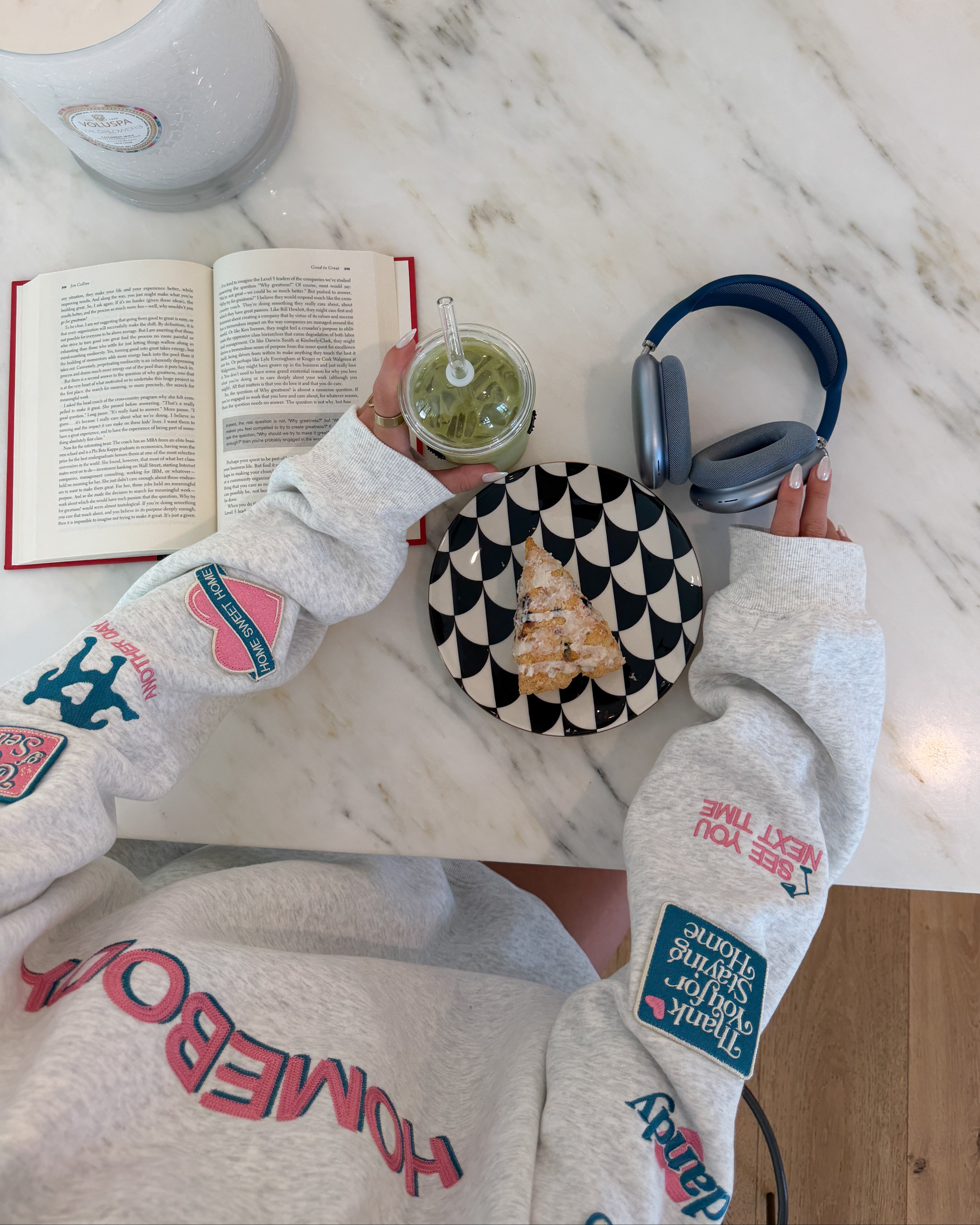 Person wearing a sweatshirt with text, holding a drink, book, and headphones on a marble surface.