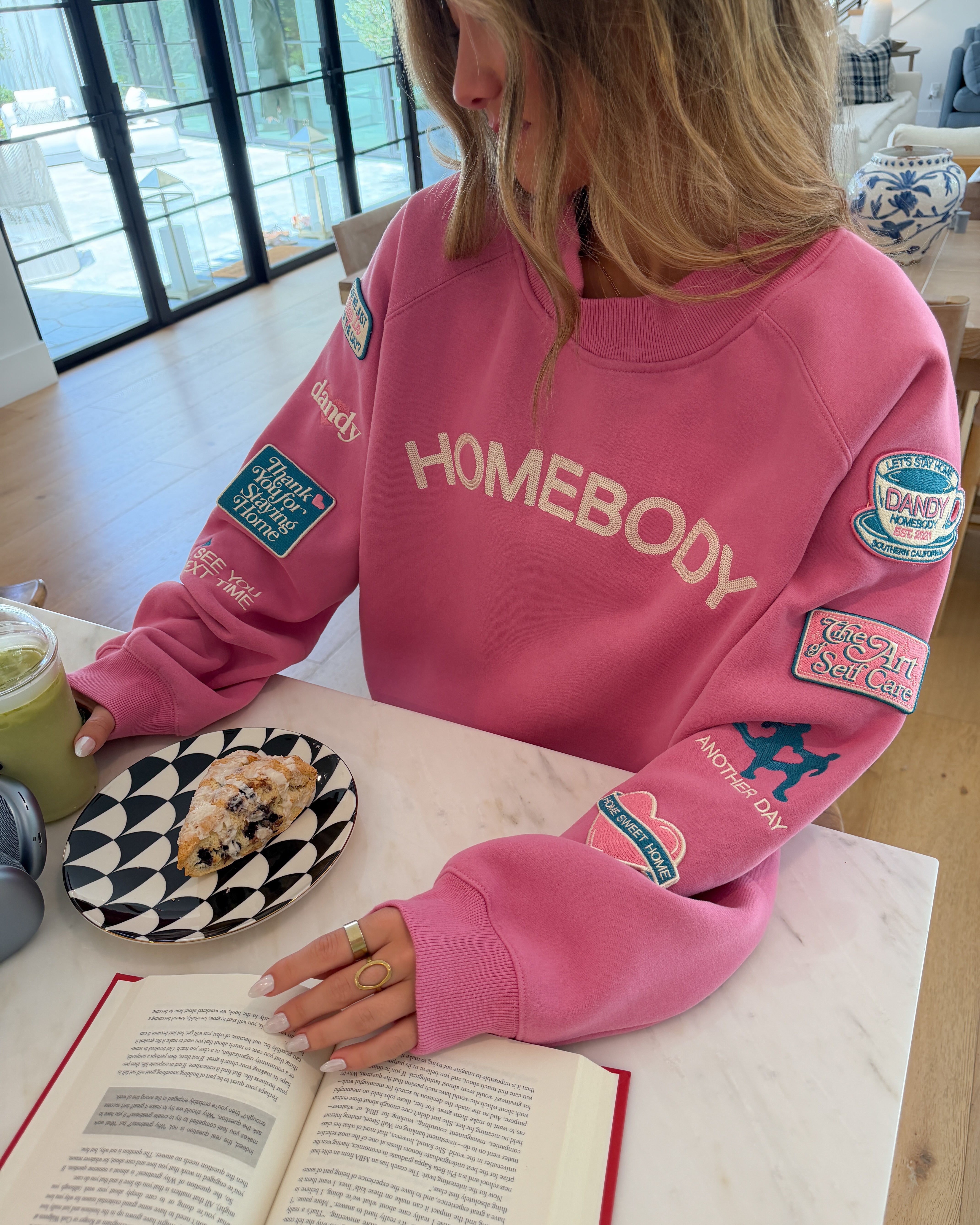 Person wearing a pink 'Homebody' sweatshirt with patches, sitting at a table with an open book and a plate.