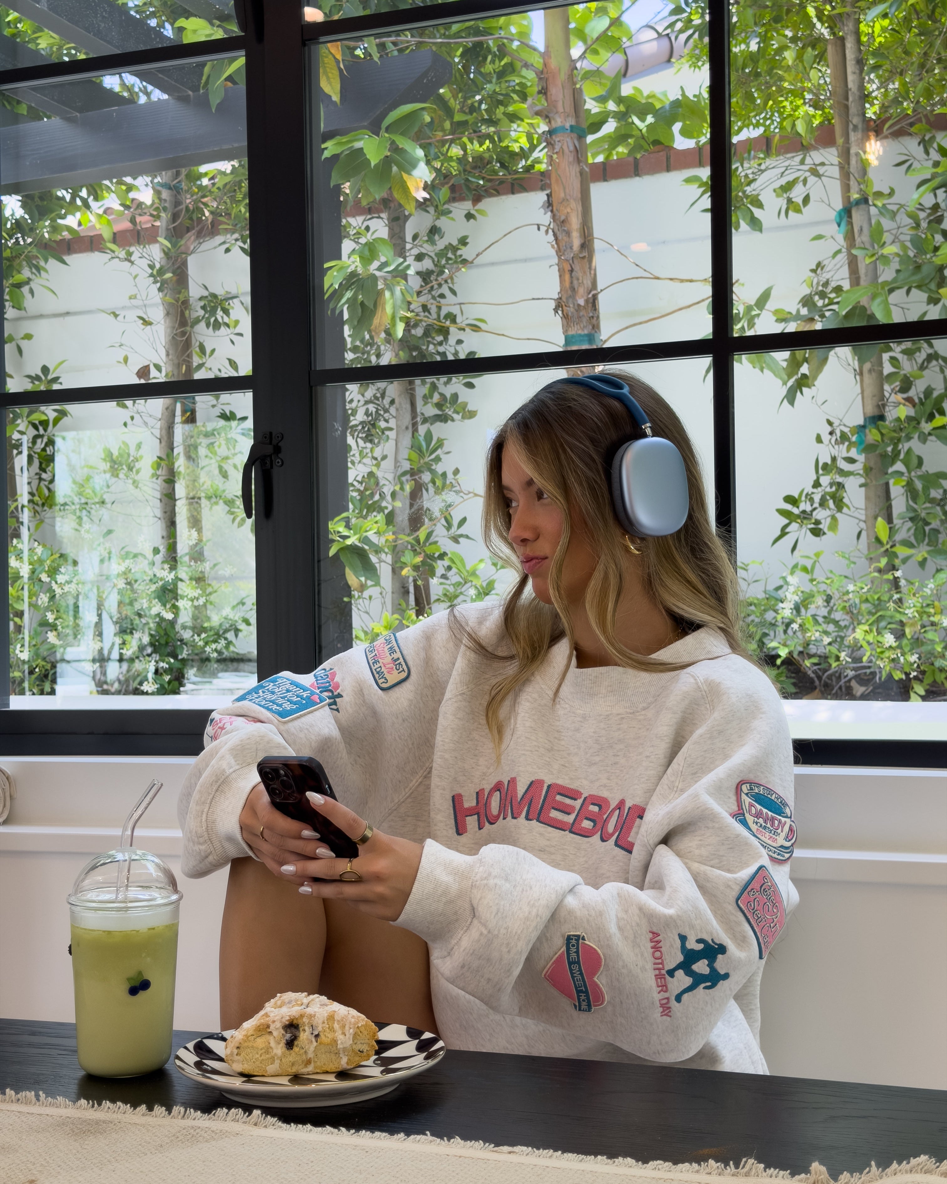 Person wearing a 'Homebody' sweatshirt sitting at a table with a drink and food, using a phone and wearing headphones.