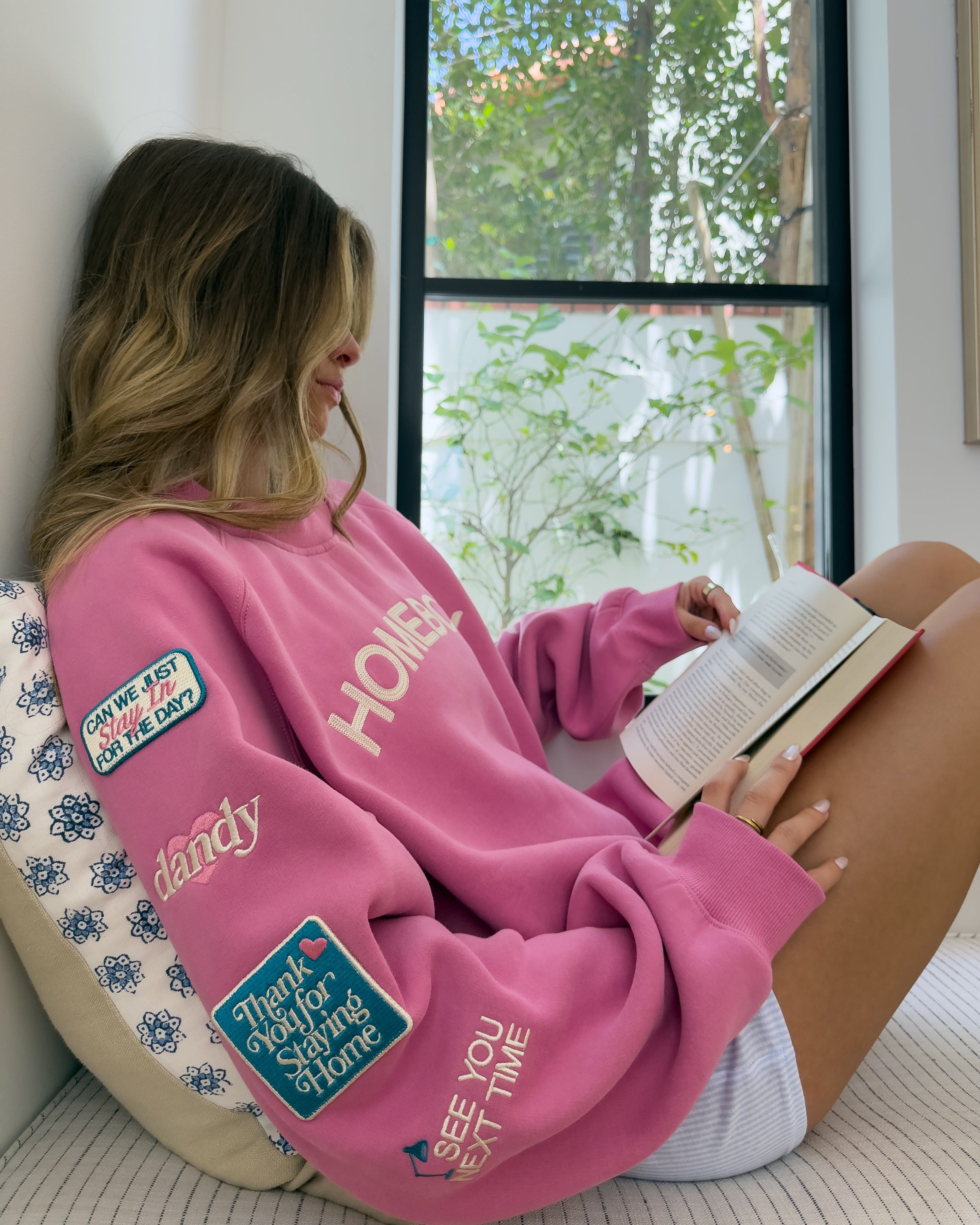 Woman in a pink sweatshirt with patches reading a book by a window.
