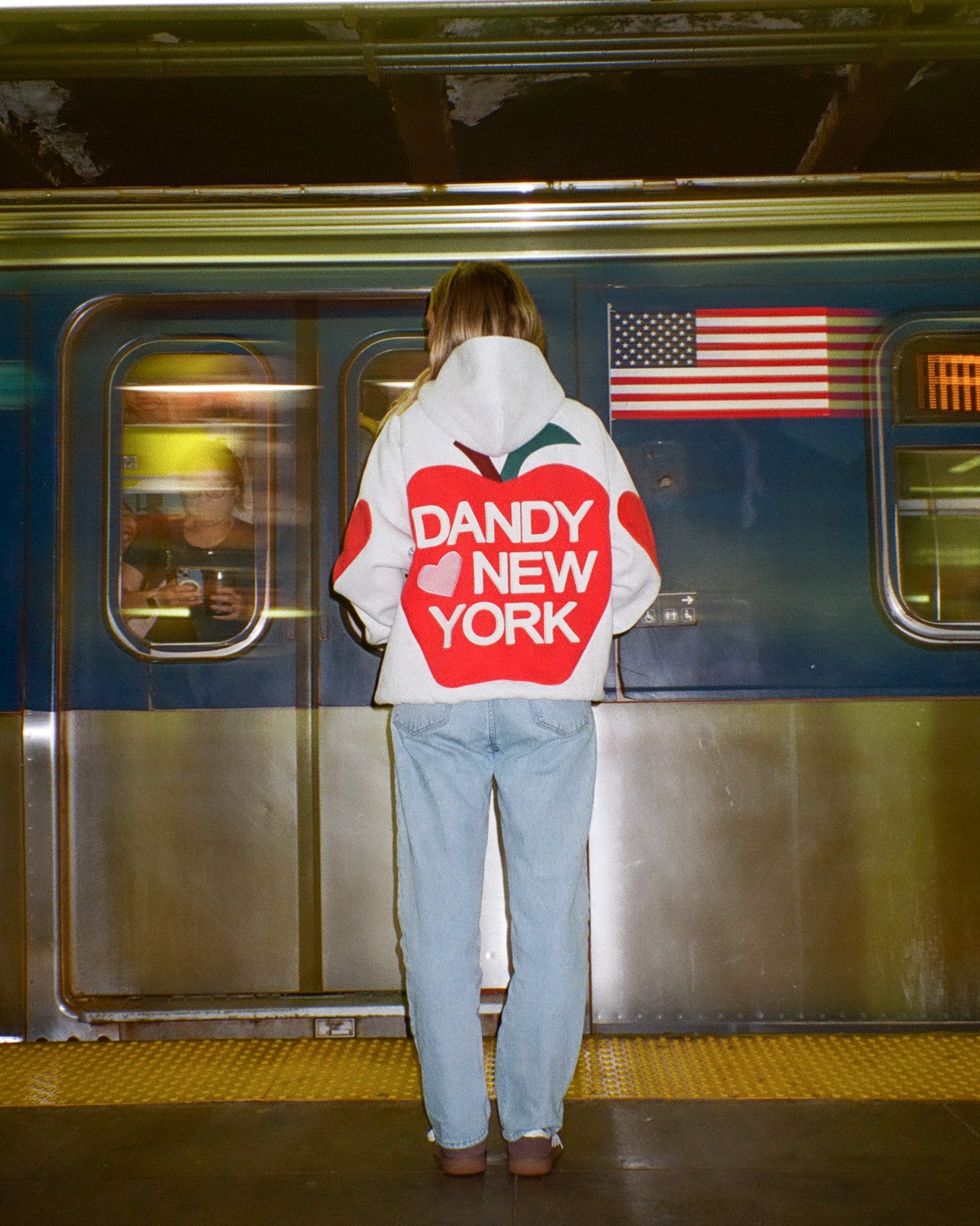 Person wearing a hoodie with 'Dandy New York' logo in front of a subway train.
