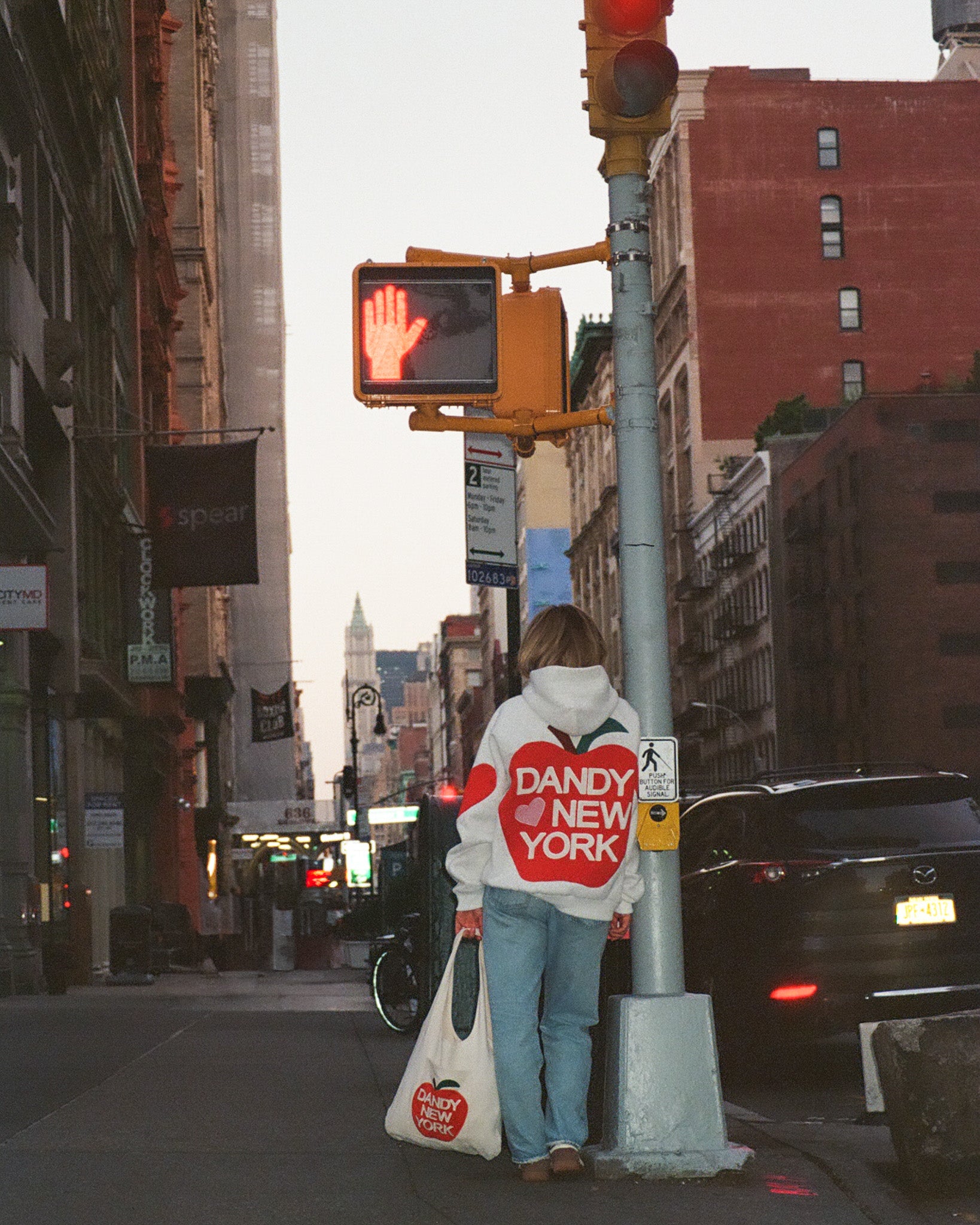 Person wearing a 'Dandy New York' hoodie standing on a city street corner with traffic lights and buildings in the background.