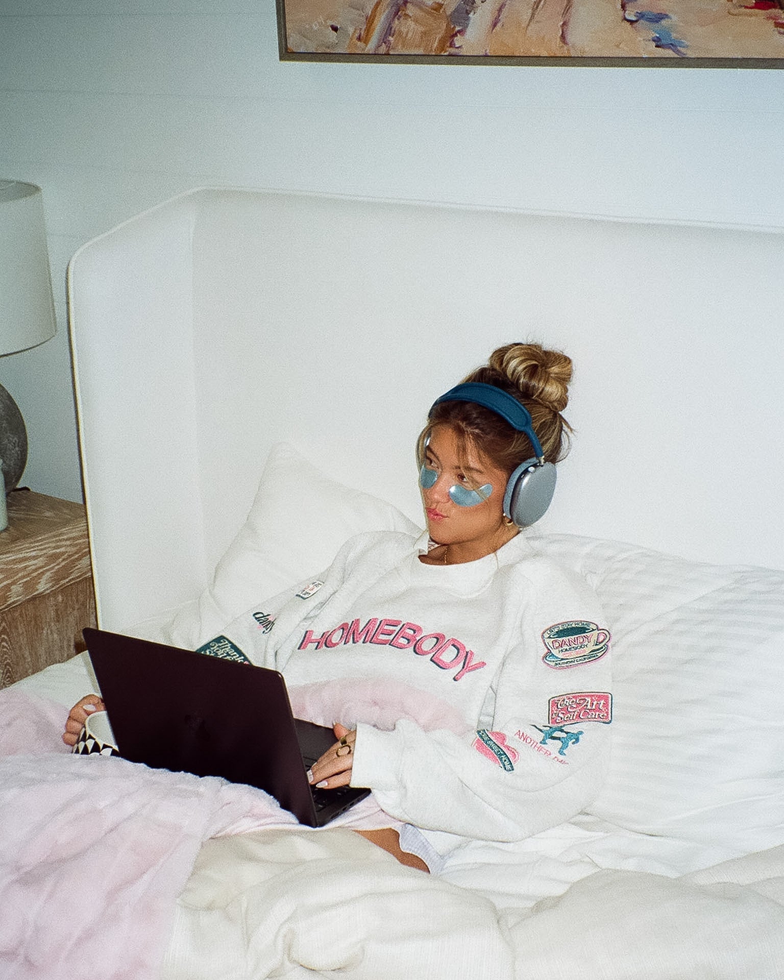Person sitting on a bed using a laptop with headphones, wearing a 'Homebody' sweatshirt.