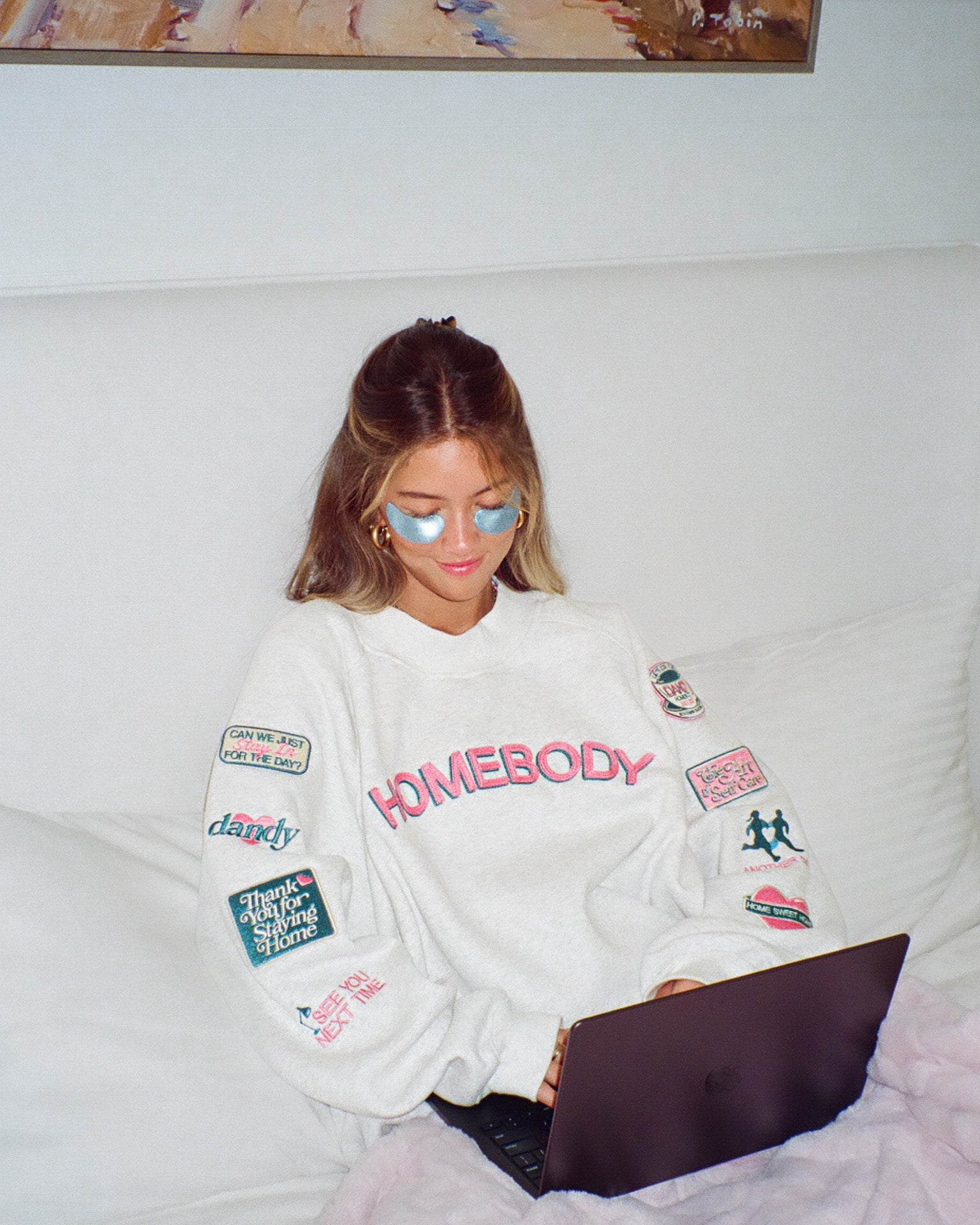 Person wearing a 'Homebody' sweatshirt with various patches, sitting on a couch using a laptop.