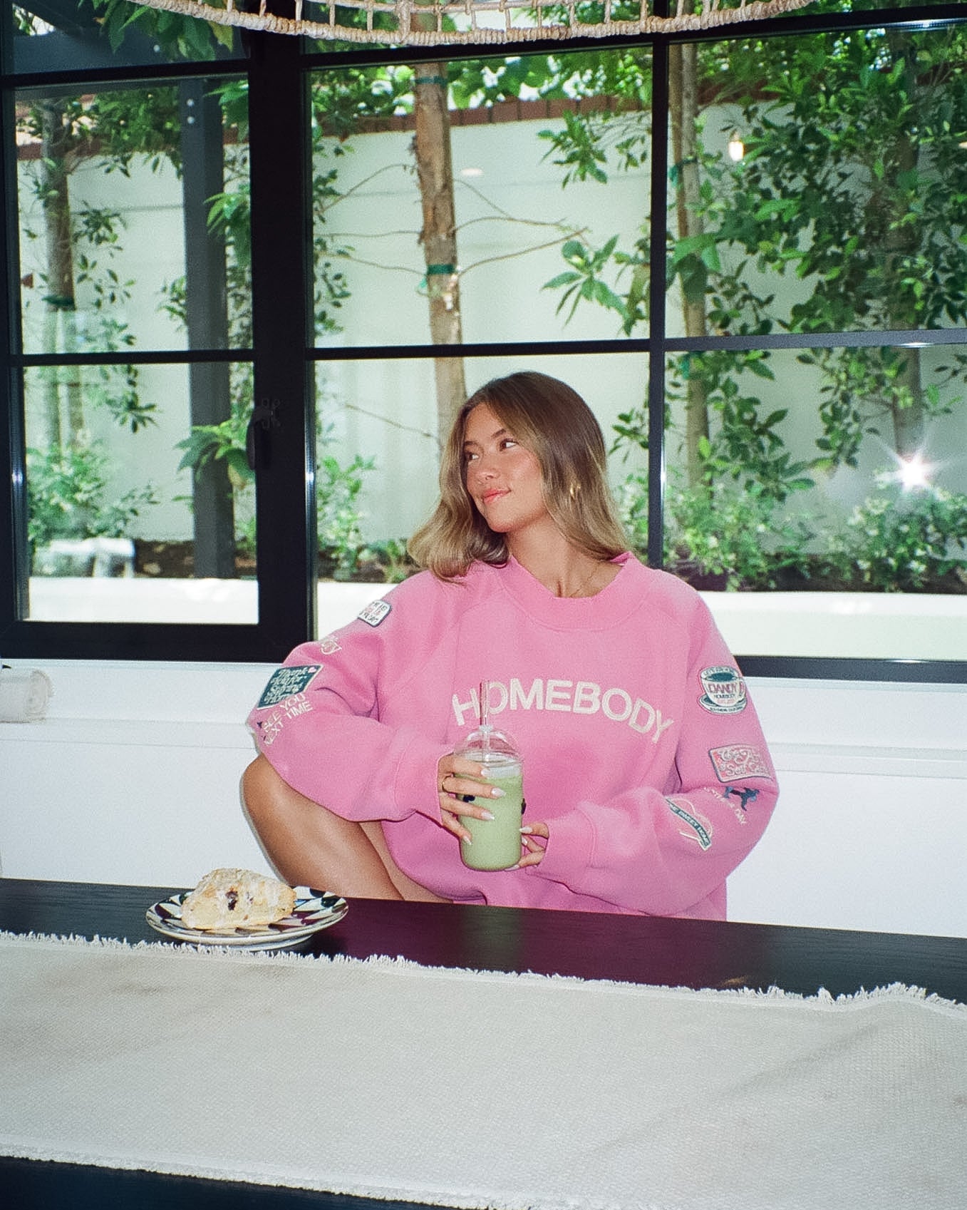 Person wearing a pink sweatshirt with 'Homebody' text, sitting at a table with a drink and dessert.